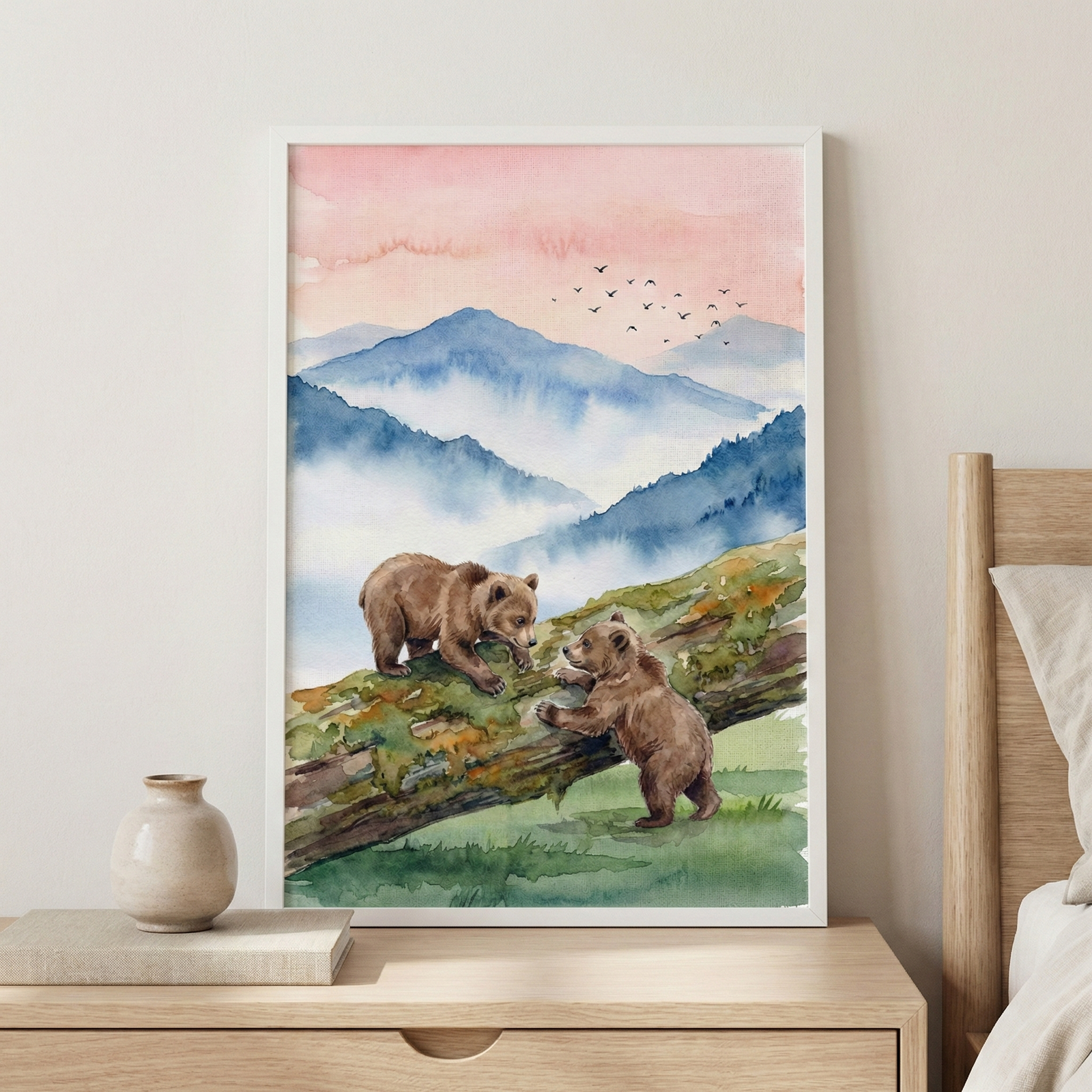 A framed watercolor illustration depicting two brown bear cubs playing on a mossy log in a mountainous landscape. The scene features soft, layered blue mountains appearing through mist, set against a peach-colored sky with a flock of birds flying in the distance. The artwork is displayed in a white frame on a light-colored wall above a wooden nightstand.