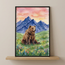 A vertical artwork in a black frame sits on a light wooden shelf. The illustration, created in a soft watercolor style, shows a large brown bear resting in a field of green grass dotted with small red, blue, and yellow wildflowers. Behind the bear, a series of dark blue mountain peaks rise toward a warm sky colored with shades of peach, pink, and pale yellow. The soft textures and blended colors give the scene a peaceful and natural feel.