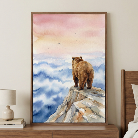 A framed watercolor illustration shows a brown bear standing on the edge of a rocky mountain peak, viewed from behind. The bear looks out across an endless layer of soft blue and white clouds. The sky above is a gentle wash of pink, orange, and yellow hues, with a few small birds flying in the distance. The artwork, characterized by fluid brushstrokes and soft color transitions, is housed in a thin wooden frame.