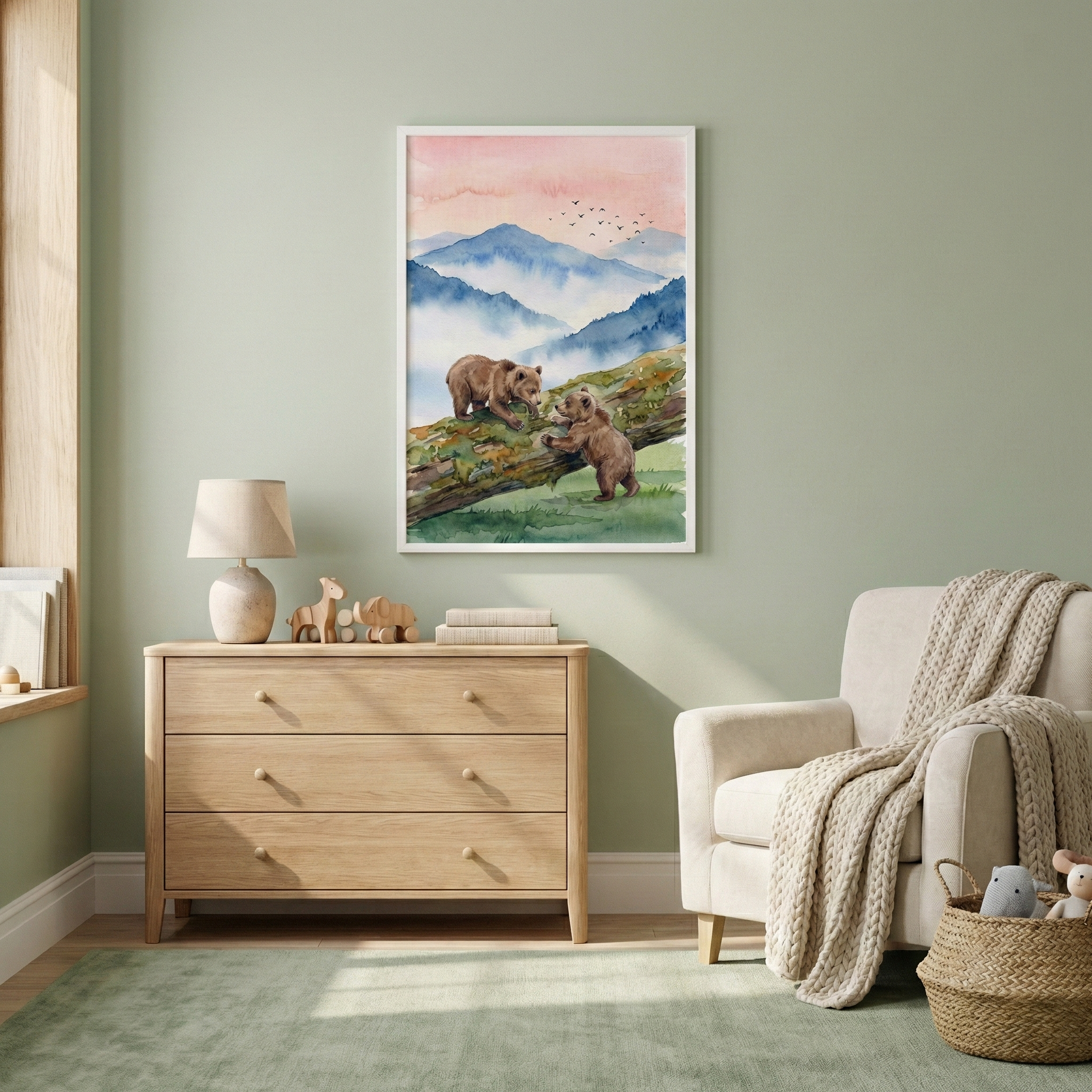 A tranquil nursery features sage green walls and a light-colored wooden floor with a soft green rug. Centered on the wall is a white-framed watercolor illustration depicting two brown bears on a fallen log amidst a backdrop of blue mountains and a pale pink sky. Below the art sits a three-drawer light wood dresser adorned with a lamp, books, and wooden animal toys. To the right, a cream armchair is draped with a textured throw, next to a woven basket filled with plush toys.