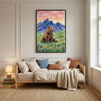 A framed vertical watercolor illustration of a brown bear sitting in a meadow of wildflowers. Behind the bear are blue mountains and a pink and orange sunset sky. The art is centered on a beige wall above a wooden daybed. The daybed is styled with pillows in shades of white, blue, and rust, and a chunky knit throw. To the left, a small lamp glows on a stack of books. A woven rug is on the floor, and a window to the right lets in natural light.