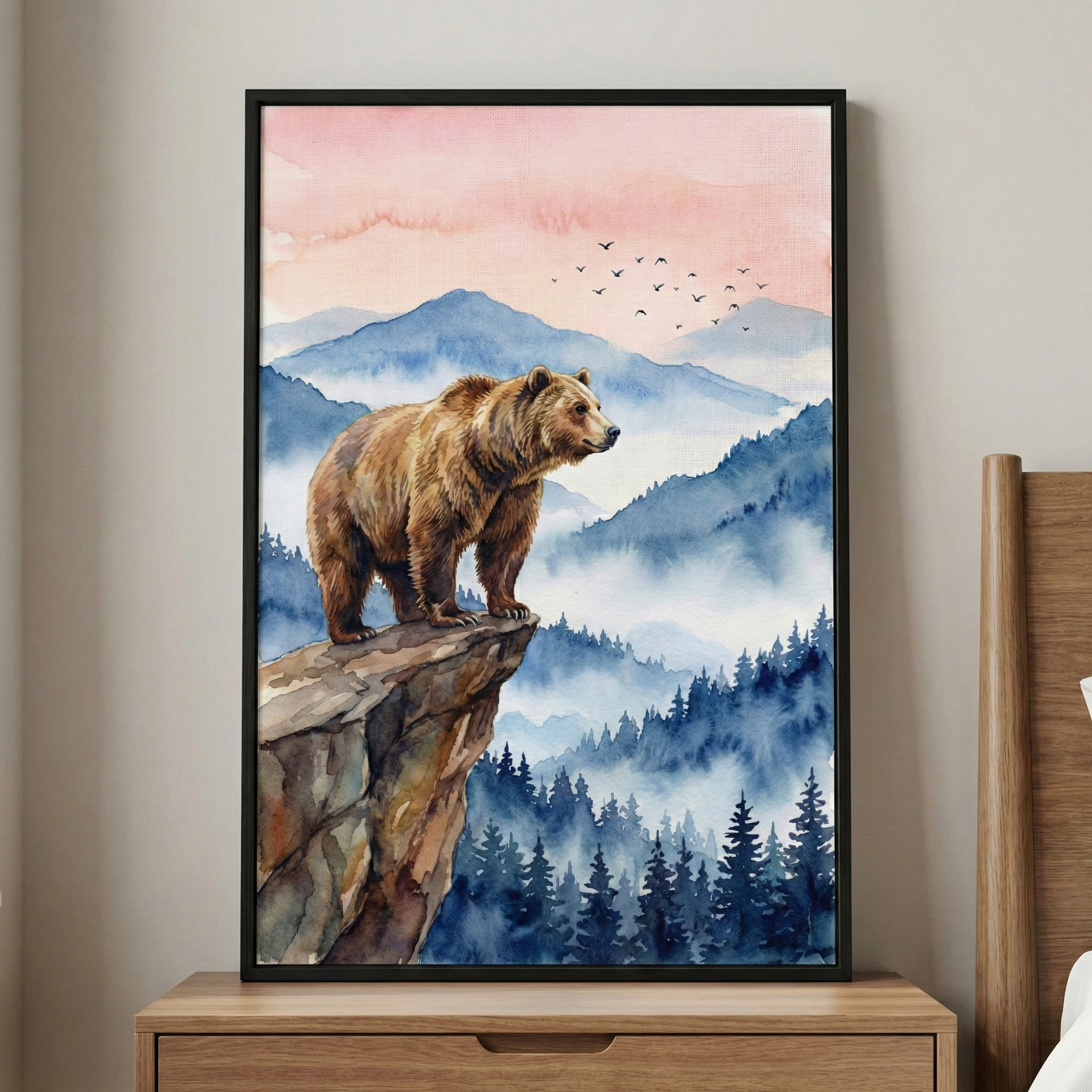 A framed watercolor print shows a brown bear standing on the edge of a rocky cliff overlooking misty blue mountains and a dense pine forest below. The sky fades from soft peach to pale pink with a small flock of birds in flight, creating a peaceful woodland sunrise scene. The artwork sits on a wooden dresser against a light neutral wall, adding a calm nature inspired focal point to the room.