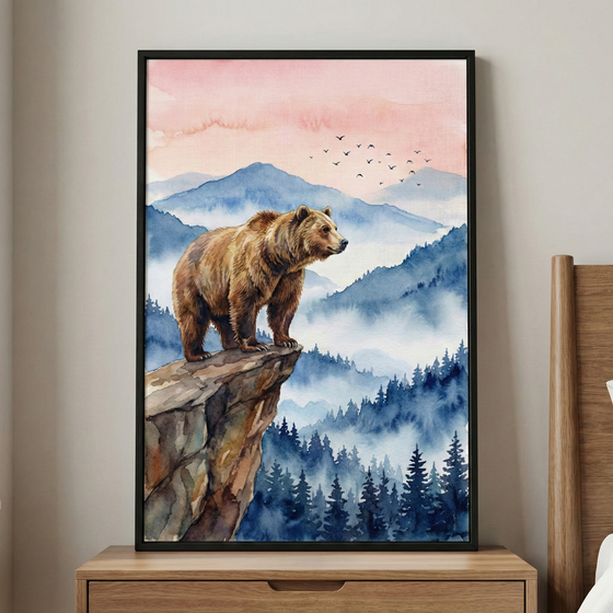 A framed watercolor print shows a brown bear standing on the edge of a rocky cliff overlooking misty blue mountains and a dense pine forest below. The sky fades from soft peach to pale pink with a small flock of birds in flight, creating a peaceful woodland sunrise scene. The artwork sits on a wooden dresser against a light neutral wall, adding a calm nature inspired focal point to the room.