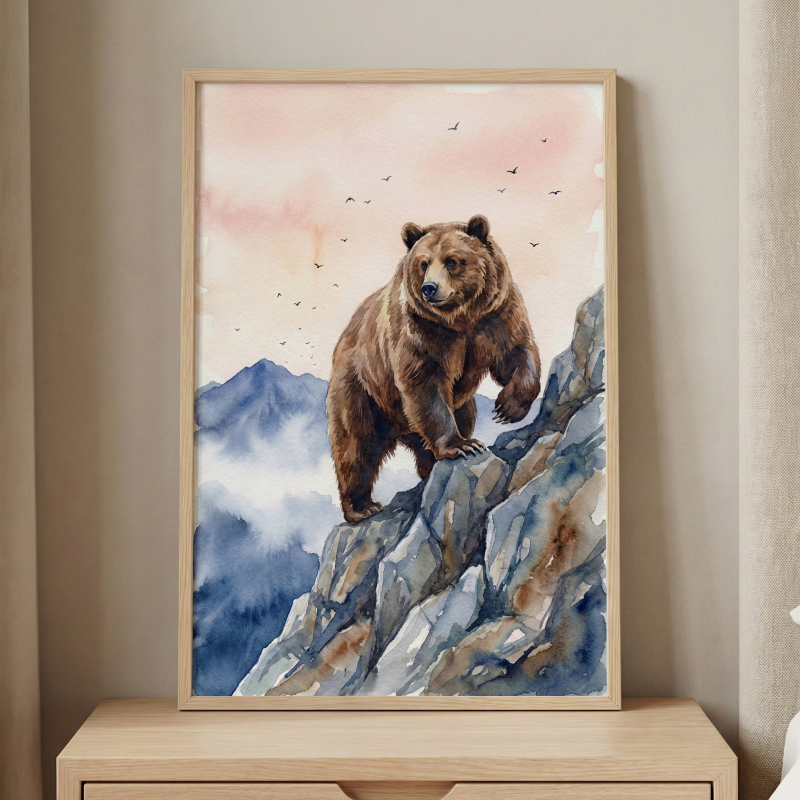 A framed watercolor-style artwork depicting a large brown bear standing on a craggy, blue-toned rocky mountain ledge. In the background, misty blue mountain peaks rise under a soft sky colored with pale pink and orange hues. A small flock of birds is silhouetted against the sky. The art is set within a light-colored wood frame, resting on a wooden surface against a neutral-toned wall.