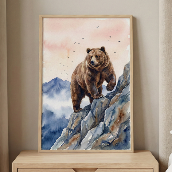 A framed watercolor-style artwork depicting a large brown bear standing on a craggy, blue-toned rocky mountain ledge. In the background, misty blue mountain peaks rise under a soft sky colored with pale pink and orange hues. A small flock of birds is silhouetted against the sky. The art is set within a light-colored wood frame, resting on a wooden surface against a neutral-toned wall.