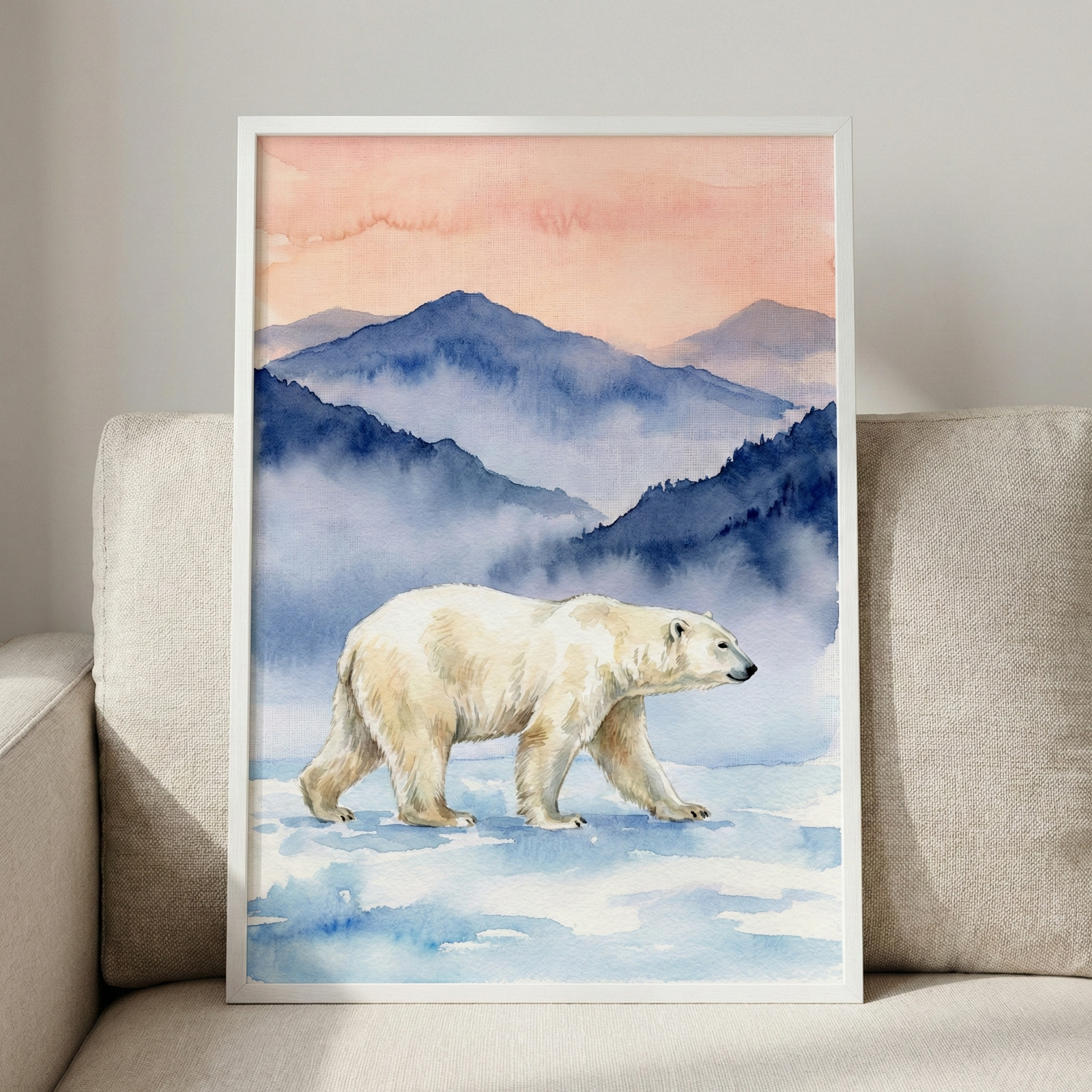 A white-framed artwork rests on a light beige fabric couch. The piece illustrates a large polar bear walking across a snowy, icy surface. Behind the bear, several layers of hazy blue mountains fade into the distance, partially obscured by mist. The sky above is rendered in soft washes of peach and warm orange, suggesting a sunrise or sunset. The entire scene is executed in a delicate watercolor style with fluid textures and soft transitions. 