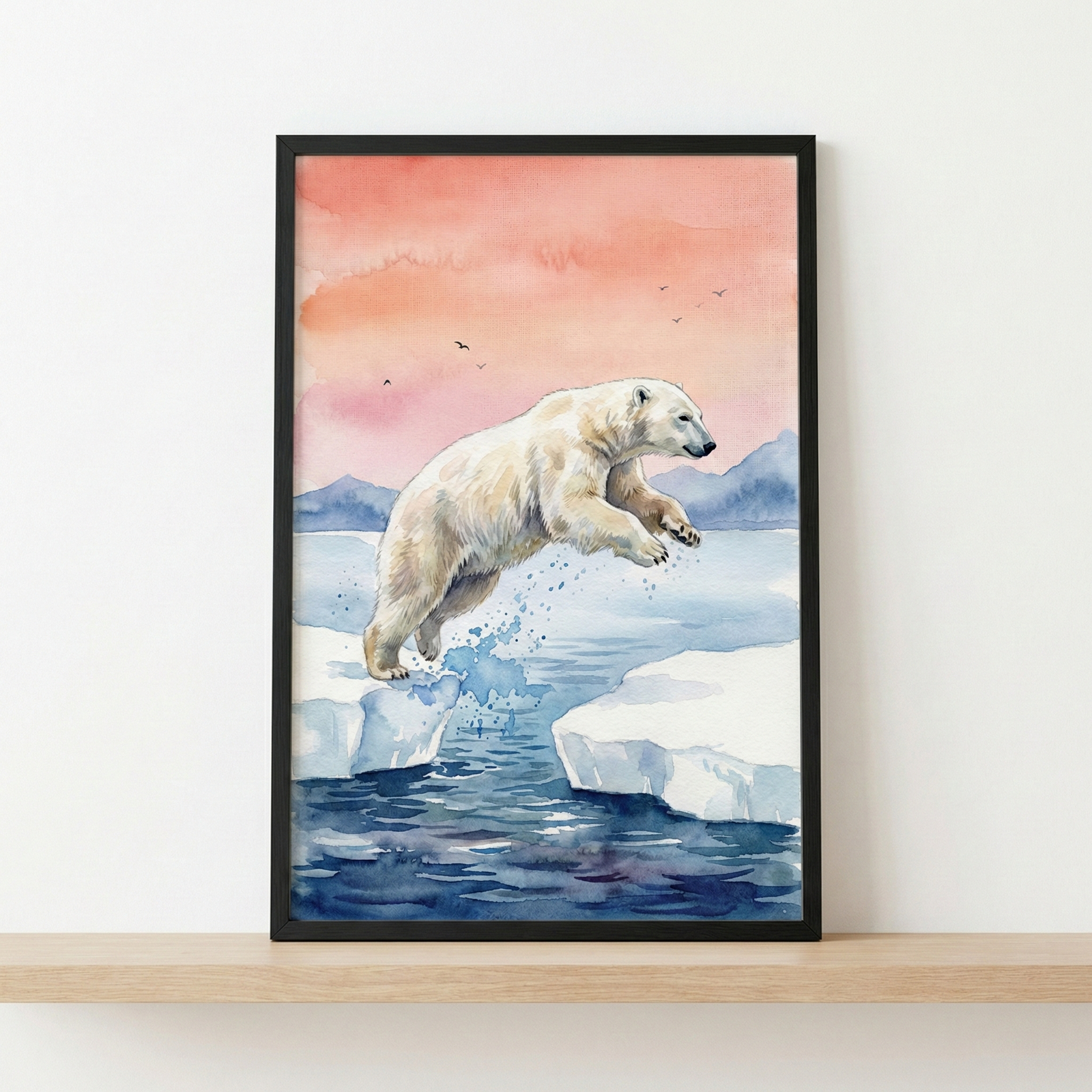 A framed watercolor illustration of a polar bear jumping across a gap between two ice floes. The artwork sits on a light wooden shelf against a plain white wall. The scene captures the bear in mid-leap above deep blue water, with splashes between the ice. In the background, jagged blue mountains sit under a soft sky with washes of pink and peach, dotted with small bird silhouettes. The artwork is presented in a simple, thin black frame.