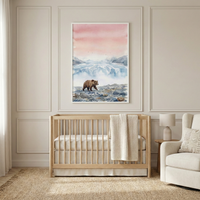 An elegant nursery scene features a framed watercolor artwork as its centerpiece. The illustration portrays a lone brown bear traversing a rocky coastline. Behind the animal, a vast blue and white glacier stretches distant mountains under a tranquil, pink-hued sky. The piece is housed in a minimalist white frame and hangs above a light-toned wooden crib. The room's decor is completed with neutral walls accented by rectangular molding, a plush white armchair with a textured cushion, and a natural woven rug.