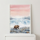 This framed watercolor illustration features a brown bear walking across a rocky shoreline. In the background, a massive blue and white glacier sits below distant, snow-capped mountains. The top half of the piece is filled with a soft, atmospheric sky rendered in a gradient of warm pink and coral tones, with tiny birds flying in the distance. The artwork is housed in a thin white frame and displayed on a sleek white surface against a minimalist wall.