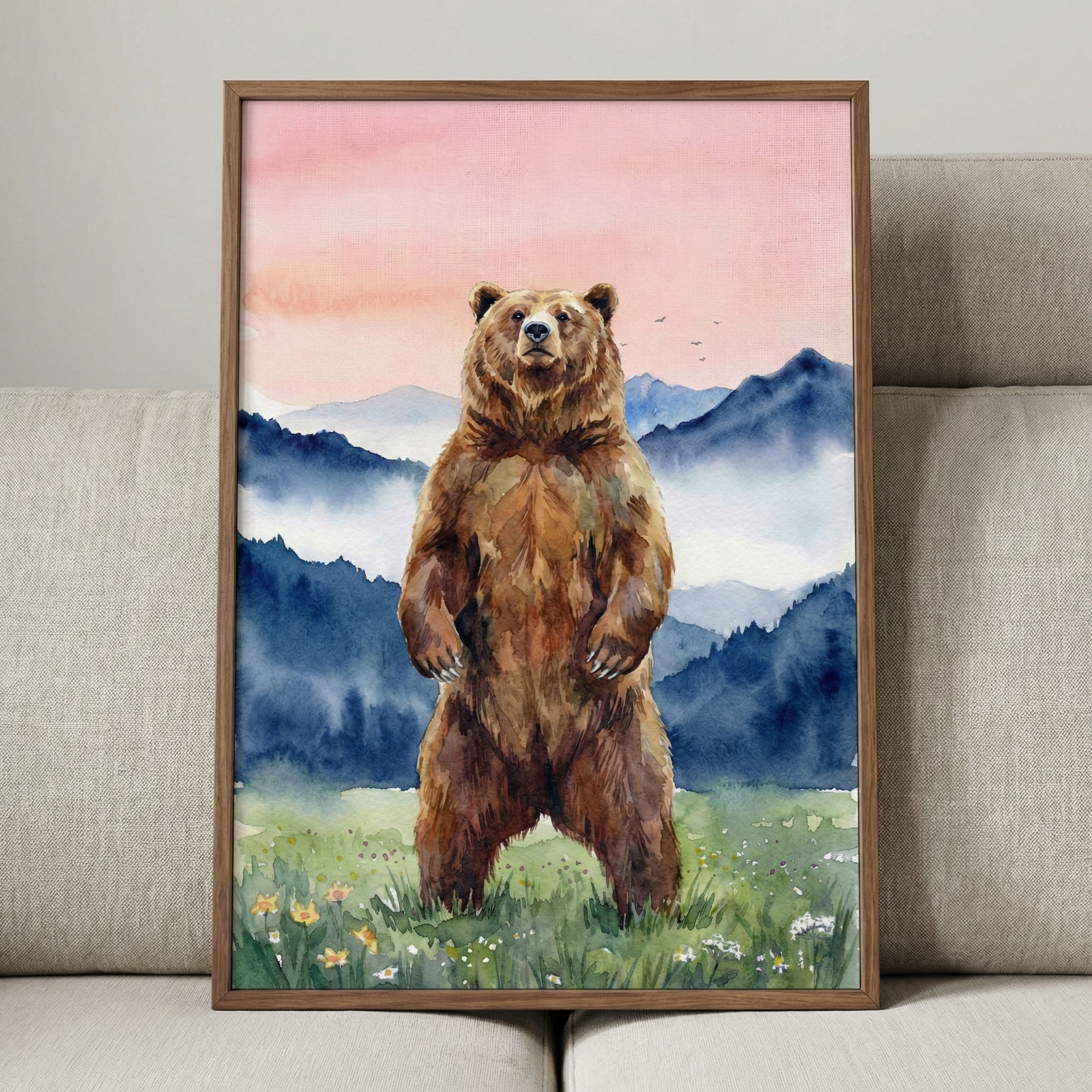 A vertical watercolor artwork featuring a large brown bear standing tall on its hind legs in a grassy meadow dotted with small yellow and white flowers. Behind the bear, layers of mist-covered mountains transition from dark blue to soft indigo. The sky above is a delicate blend of pink and peach colors, with a few small bird silhouettes flying in the distance. The piece is enclosed in a simple brown wood frame and is shown leaning against a light grey textured sofa.