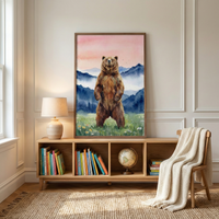 A large rectangular wooden frame hangs on a light-colored wall with classic architectural molding. The artwork inside features a brown bear standing on its hind legs in a meadow filled with small wildflowers. The background of the illustration depicts misty blue mountains beneath a soft sky with shades of pink and peach.

