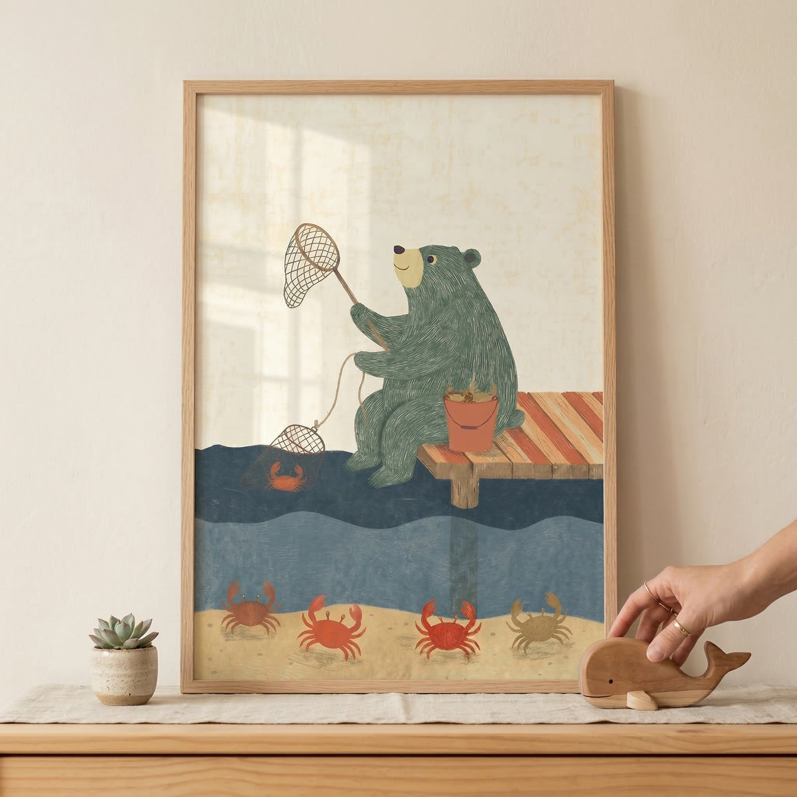 Framed children’s illustration of a green bear sitting on a wooden dock holding two crab nets above the water while red crabs walk along the sandy bottom below. The framed print rests on a dresser beside a small plant while a hand places a wooden whale toy nearby.