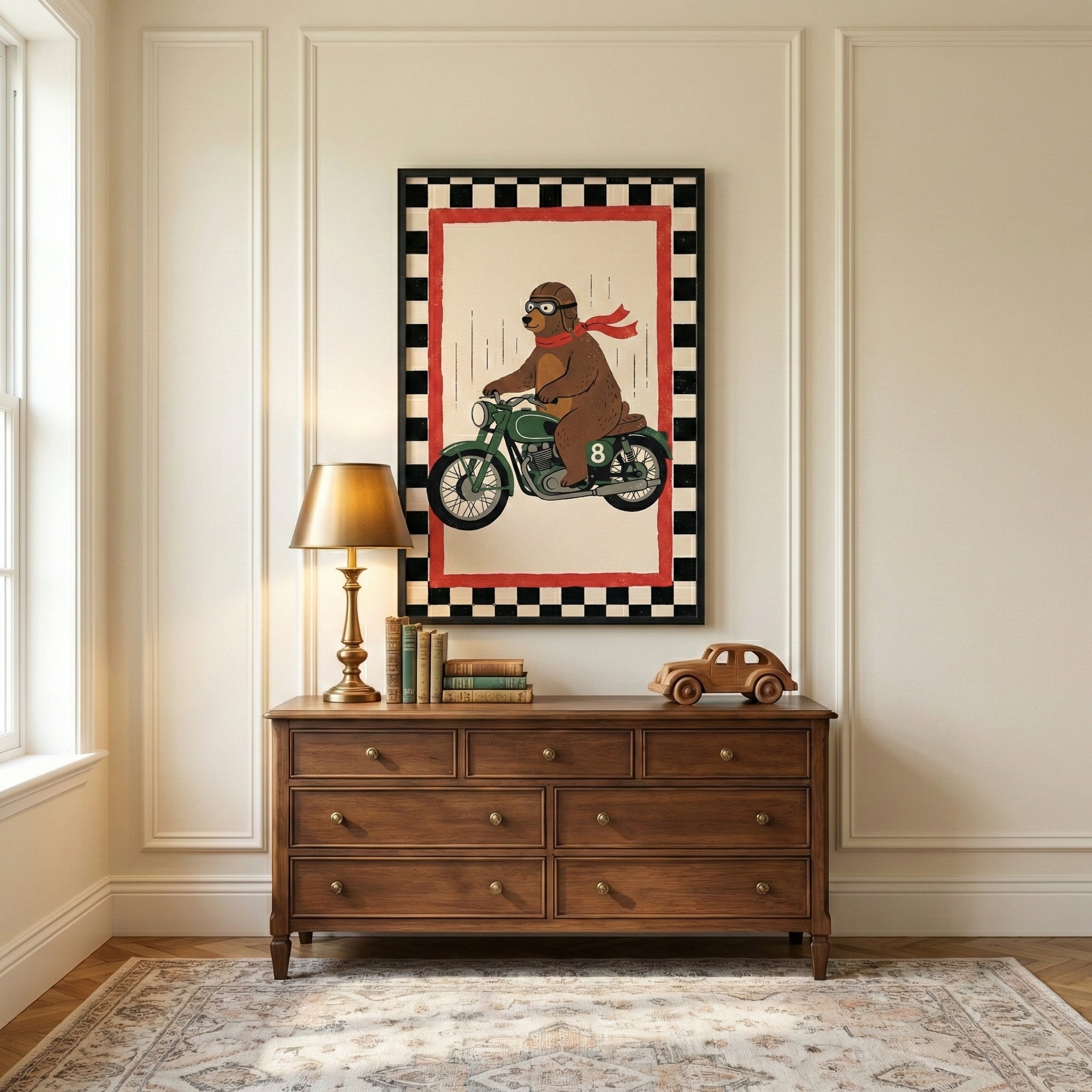 A framed illustration of a brown bear wearing goggles and a red scarf while riding a green motorcycle hangs on a cream-colored wall with decorative paneling. Below the artwork sits a wooden seven-drawer dresser. On the dresser's top surface are a gold lamp with a shade, a small stack of books, and a wooden toy car. A patterned area rug lies on the floor, and a window is partially visible on the left.