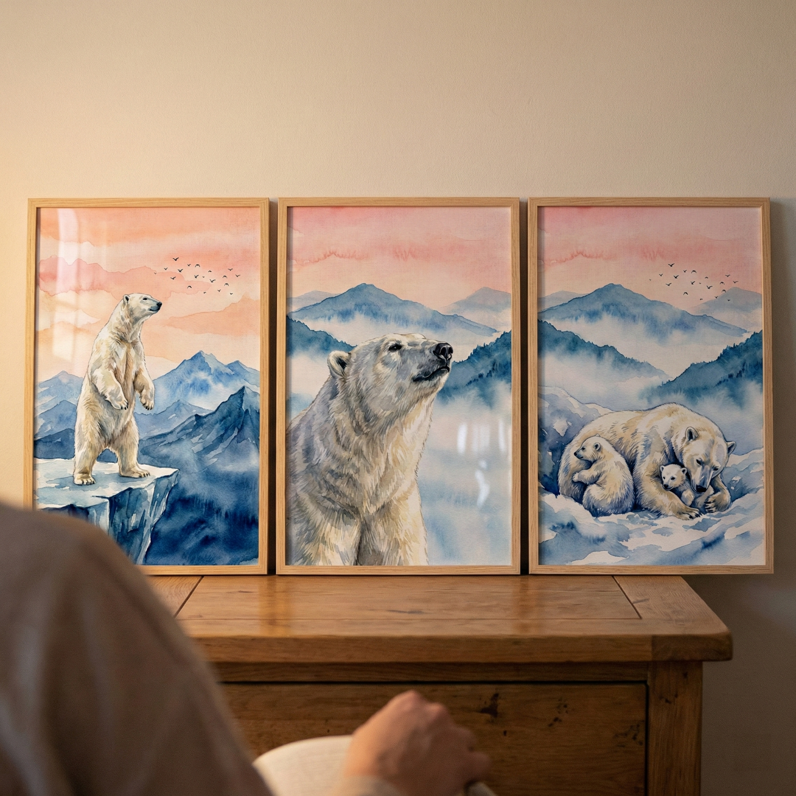 Three wooden-framed watercolor illustrations of polar bears are displayed side-by-side on a light wood surface. Each piece features a serene landscape of icy blue mountains under a soft pink and orange sunset sky. The left frame shows a polar bear standing on a jagged cliff. The center frame is a close-up portrait of a bear's face. The right frame depicts a mother bear cuddling two small cubs. A blurred figure is visible in the foreground, viewing the triptych of artwork.