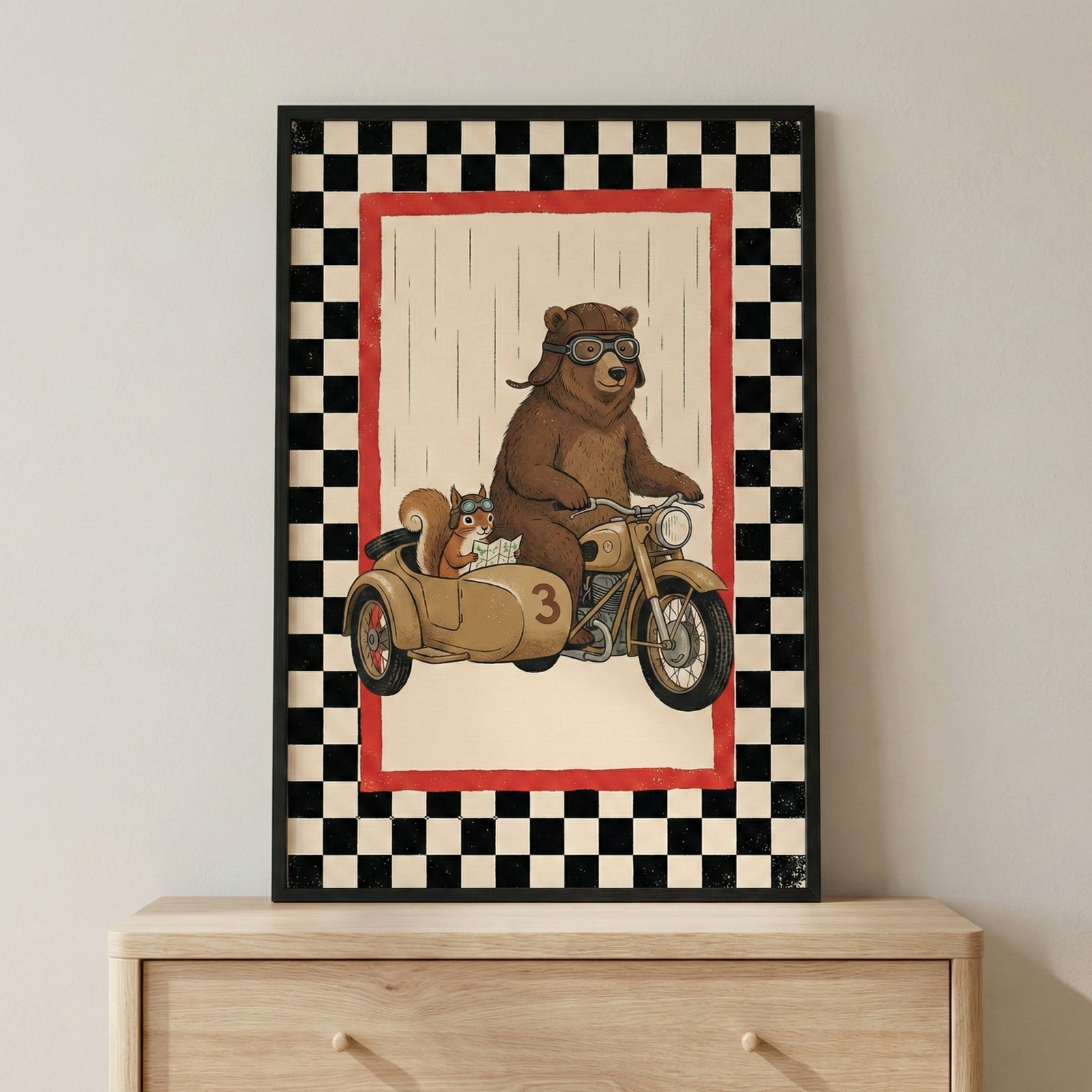 A framed illustration sits on a light wooden dresser against a neutral wall. The artwork depicts a large brown bear wearing a leather pilot's cap and goggles, riding a tan-colored vintage motorcycle. A small squirrel, also wearing goggles and holding a paper map, sits in the motorcycle's sidecar, which is marked with the number three. The central scene is set against a pale background with subtle vertical lines and is framed by a thin red border followed by a wide black-and-white checkered pattern.