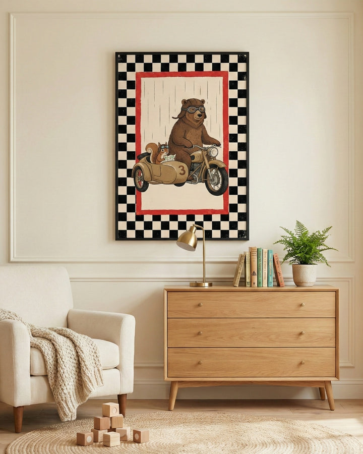 A cozy interior scene featuring a piece of framed wall art centered on a light-colored wall with elegant molding. The illustration shows a brown bear and a small squirrel, both wearing goggles, riding a vintage motorcycle with a sidecar marked with the number three. The art is finished with a bold black-and-white checkered border and a red inner frame. Underneath the art sits a three-drawer wooden dresser holding a small brass lamp, a row of books, and a green potted plant. 