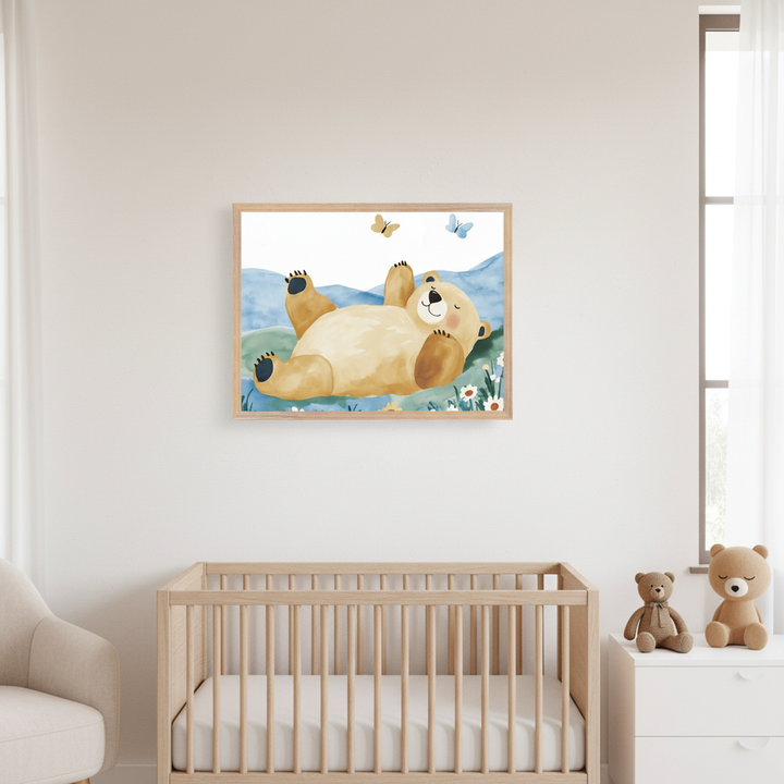 Modern nursery with a framed watercolor print of a happy light brown bear lying on its back in a field with blue hills and butterflies, hung above a natural wood crib. A white dresser with two teddy bears sits right; a beige armchair is left.