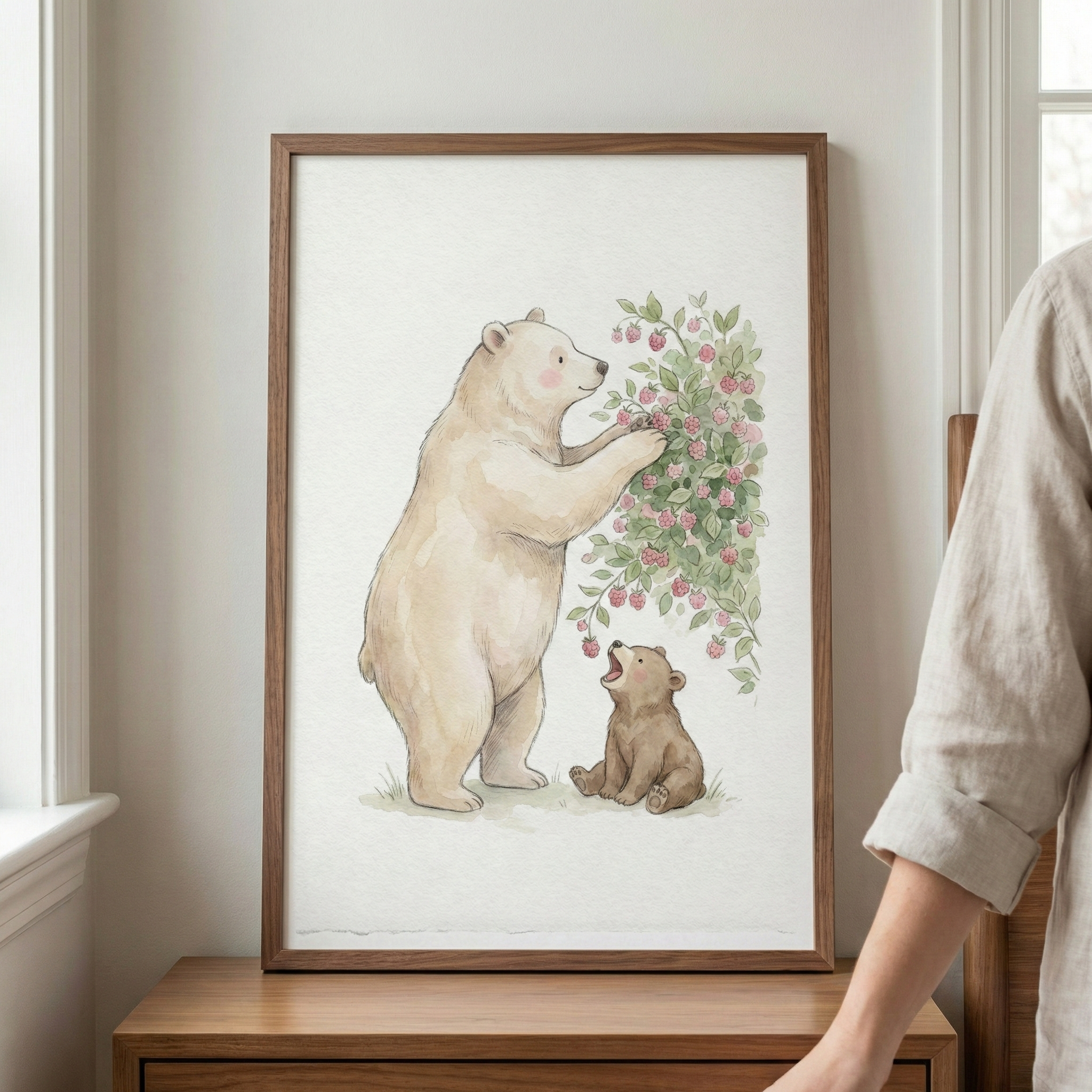 A framed watercolor illustration depicts a large, cream-colored bear standing on its hind legs to reach small pink berries on a leafy bush. Beside it, a small brown cub sits on the ground, looking up with its mouth open. The artwork is set within a simple wooden frame and displayed on a wooden surface against a neutral wall. To the right, part of a person wearing a light-colored linen shirt is visible, and to the left is a white window frame.