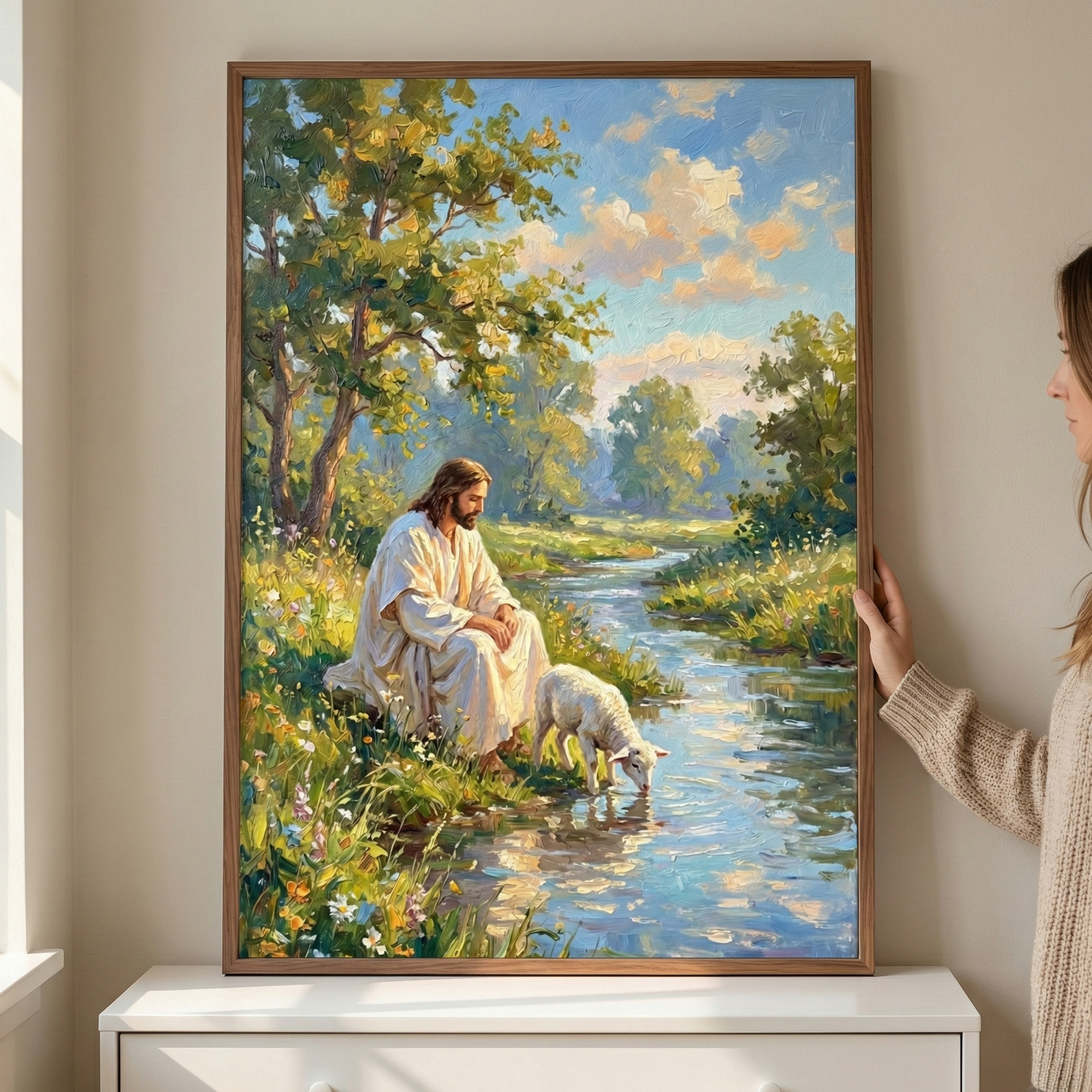 A large framed piece of art is held by a person’s hands as it rests on a white dresser. The artwork depicts Jesus in a white robe, sitting peacefully on a grassy bank filled with colorful wildflowers. Beside him, a small white lamb drinks from a clear, flowing stream. The background features lush green trees under a bright blue sky with soft, sun-drenched clouds. The style is impressionistic, with visible, textured strokes that create a bright and serene atmosphere.
