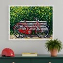 Colorful art shows a red bicycle parked beside a bench surrounded by lush greenery and bright yellow flowers. The framed art adds charm above a console table styled with a red helmet, yellow notebook, and fern, blending nature and nostalgia in a cozy setting.