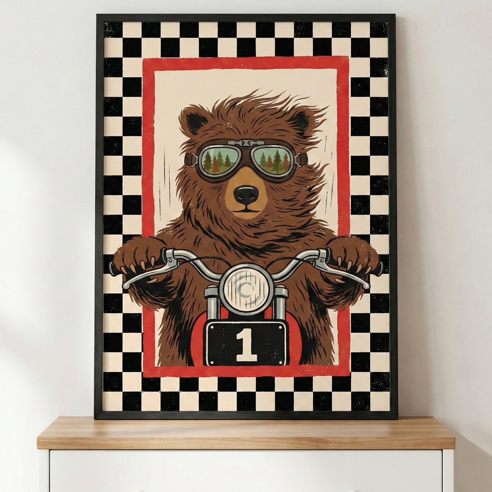 A framed illustration of a brown bear riding a motorcycle. The bear is wearing goggles that reflect a forest scene. Its paws are on the handlebars, and the motorcycle features a front headlight and a black number plate with the number one. The central image is surrounded by a red border and a wide black and white checkered pattern. The poster is in a black frame, sitting on a light-colored wooden surface.