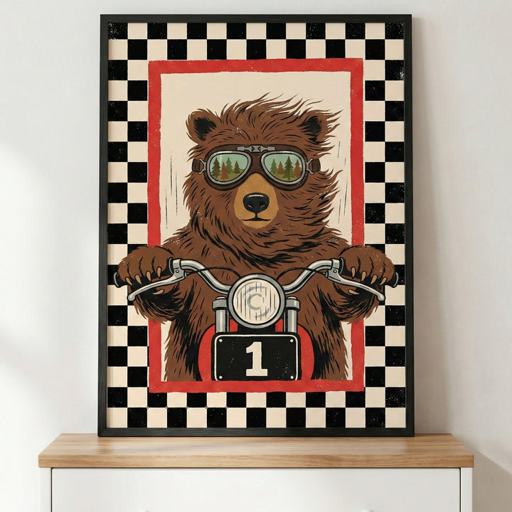 A framed illustration of a brown bear riding a motorcycle. The bear is wearing goggles that reflect a forest scene. Its paws are on the handlebars, and the motorcycle features a front headlight and a black number plate with the number one. The central image is surrounded by a red border and a wide black and white checkered pattern. The poster is in a black frame, sitting on a light-colored wooden surface.