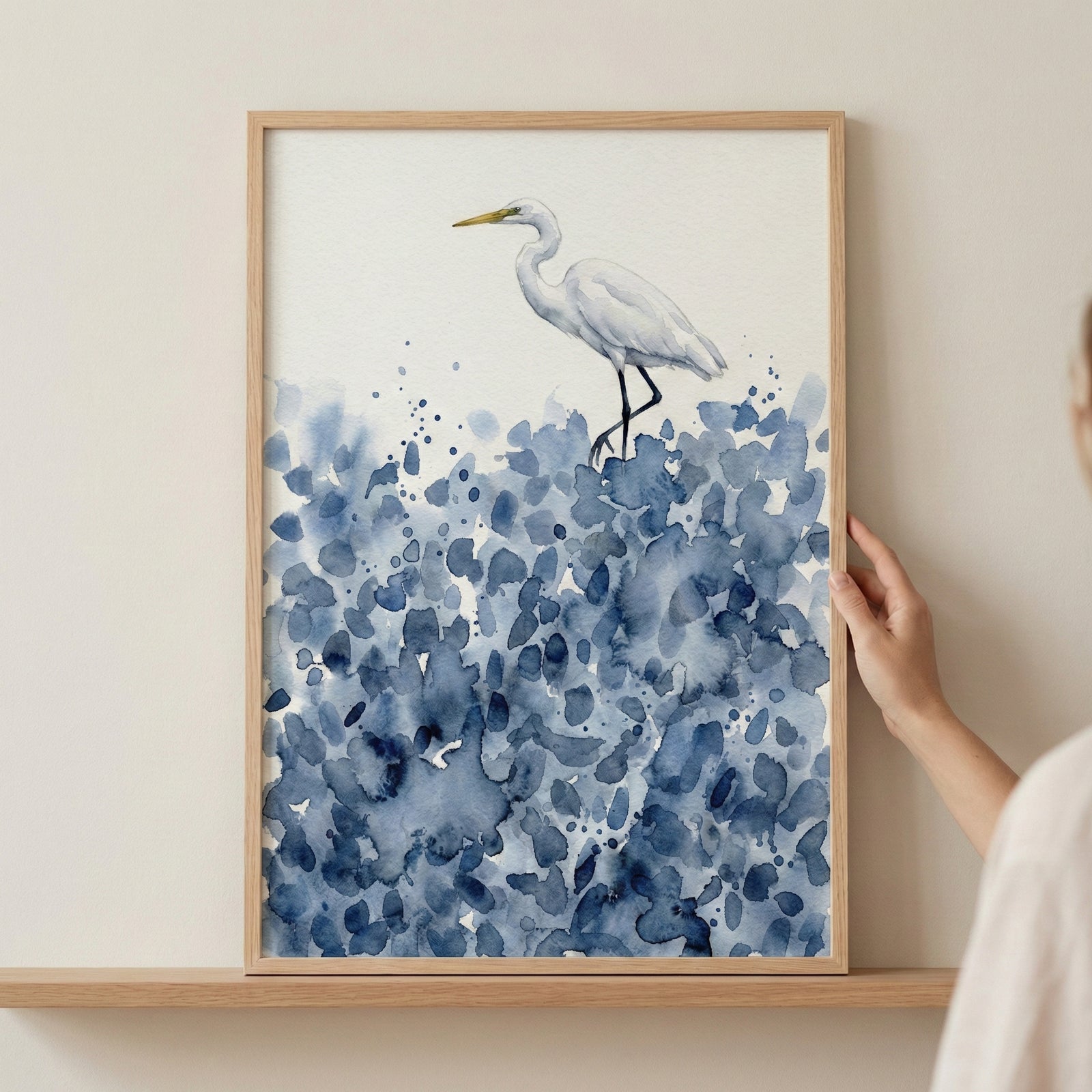 Framed watercolor painting of a white bird, likely an egret or heron, standing on a cluster of abstract blue brushstrokes resembling ocean waves or coastal foliage. The soft, minimal design evokes a peaceful nature scene.