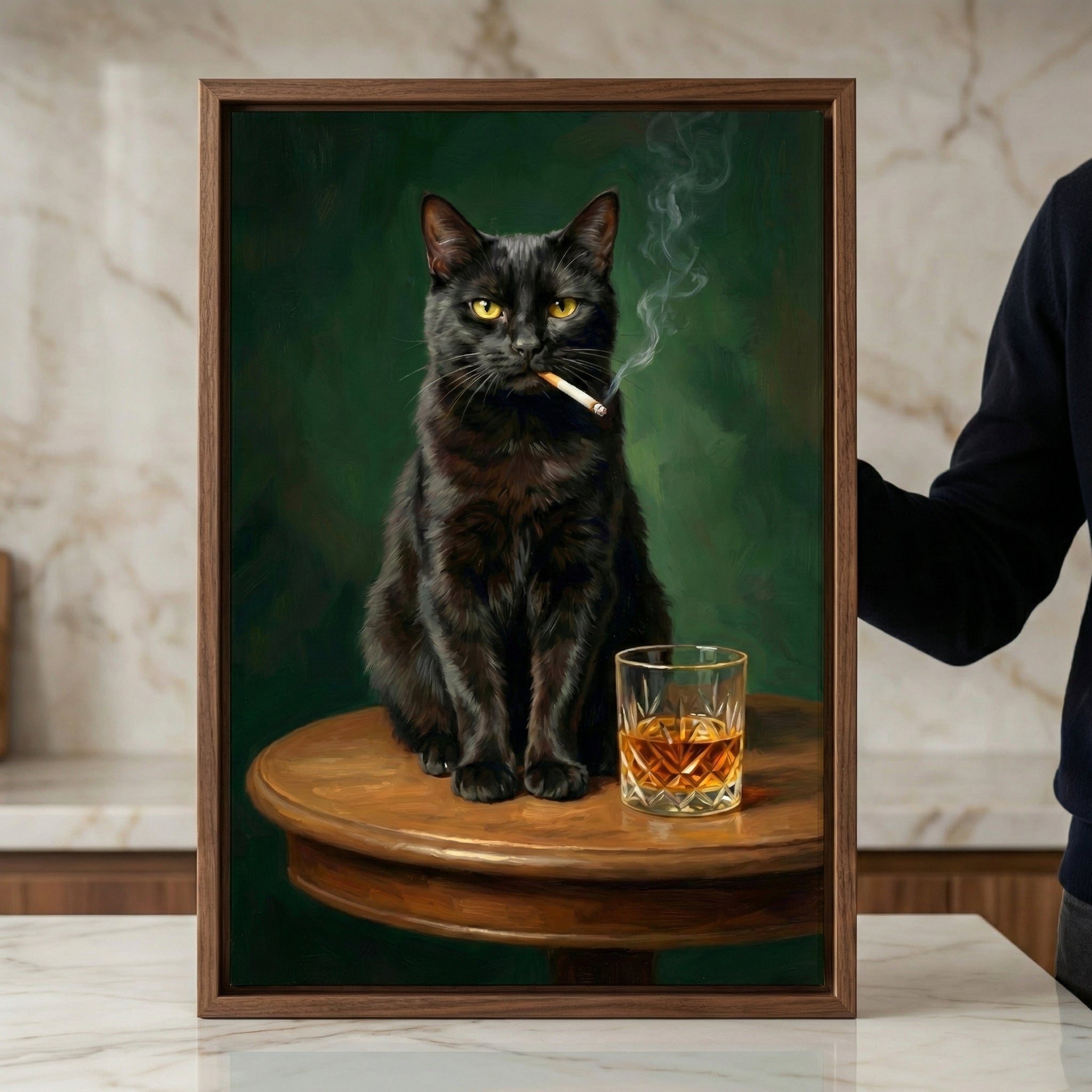 A framed vertical artwork featuring a black cat with yellow eyes sitting on a round wooden table. The cat has a lit cigarette in its mouth with thin wisps of smoke rising. Beside the cat sits a decorative glass containing an amber-colored liquid. The background is a dark, moody green. A person's arm is visible on the right, holding the wooden frame upright on a white marble surface in a modern interior setting.