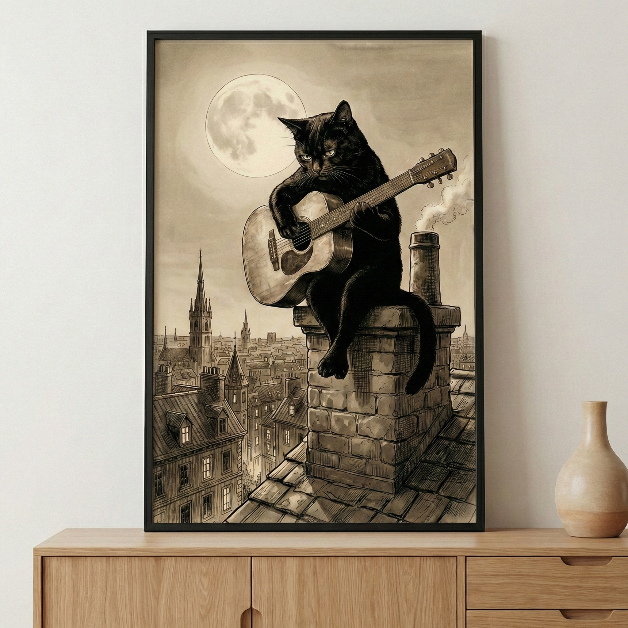 A framed sepia-toned illustration of a black cat perched on a brick chimney, playing an acoustic guitar under a large full moon. The cat sits overlooking a detailed cityscape with many spires and old-fashioned buildings. Smoke rises from a nearby chimney, adding to the nighttime atmosphere. The artwork, with its fine-line detail and vintage storybook feel, is displayed in a black frame on a wooden cabinet next to a ceramic vase.