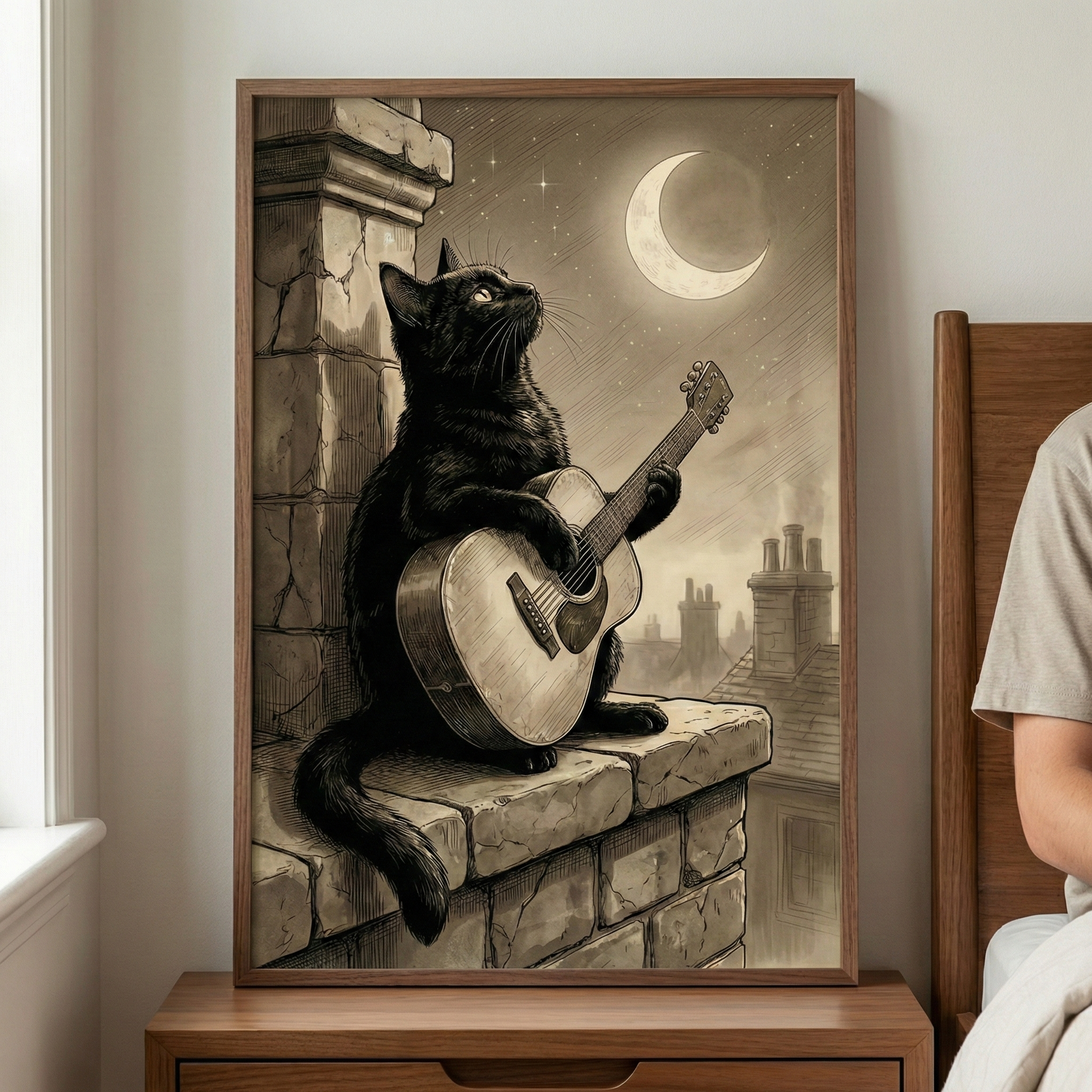 A framed illustration rests on a wooden bedside table, featuring a black cat perched on a stone ledge at night. The cat holds an acoustic guitar, looking upward toward a large, glowing crescent moon in a starry sky. Behind the cat, the silhouettes of city chimneys rise against the night air. The artwork has a vintage, sepia-toned aesthetic. Part of a person sitting on a bed is visible to the right, and a window frame appears on the left.