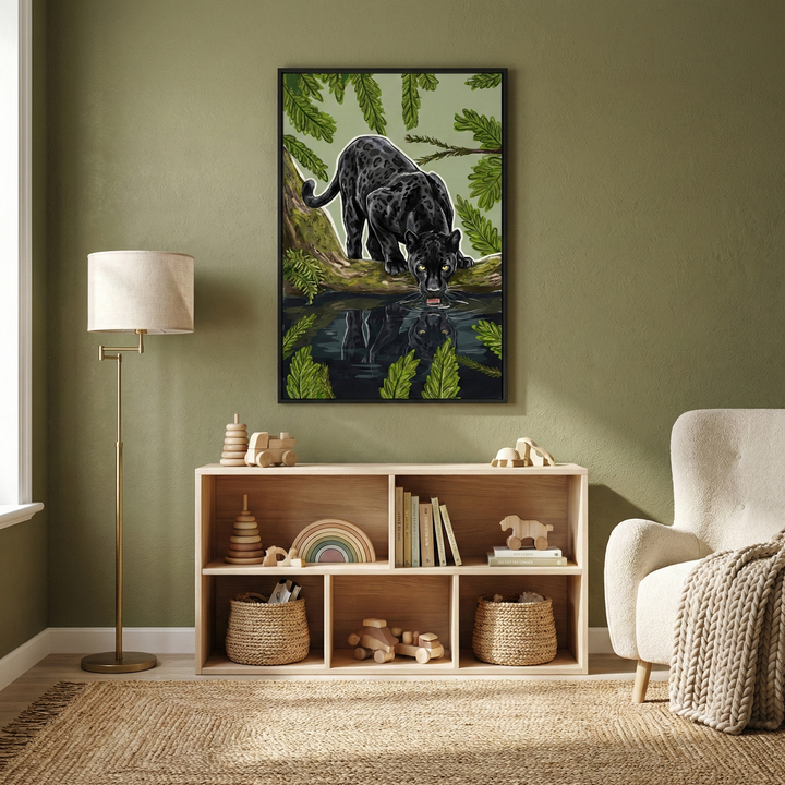 An illustrated artwork of a black panther drinking from a pool of water hangs on an olive green wall. The animal is perched on a branch, its reflection shimmering in the dark water below, surrounded by large green leaves. Below the frame is a light wood storage unit with six cubbies, holding wooden toys like stacking rings, a rainbow, and a toy horse, as well as woven baskets. A thin brass floor lamp is on the left, and a plush cream armchair with a textured throw is on the right.