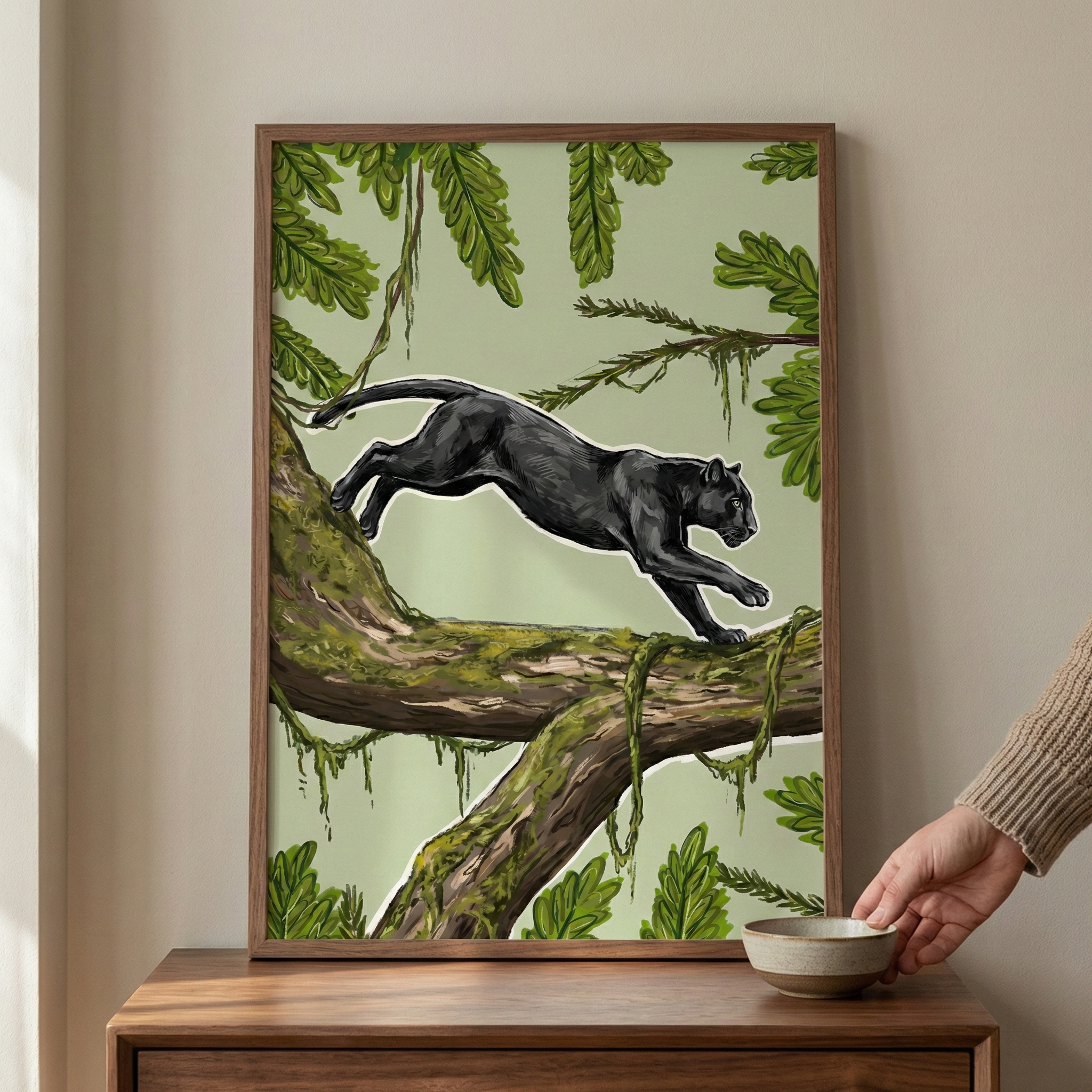 An illustration in a dark wood frame depicts a sleek black panther mid-leap across a moss-covered tree branch. Set against a pale green background, the scene is filled with lush tropical foliage, including various green leaves and hanging vines. The panther is rendered with a thin white outline that makes its form pop from the botanical surroundings. The framed piece sits on a wooden sideboard, and a hand is visible to the right, placing a small ceramic bowl nearby.