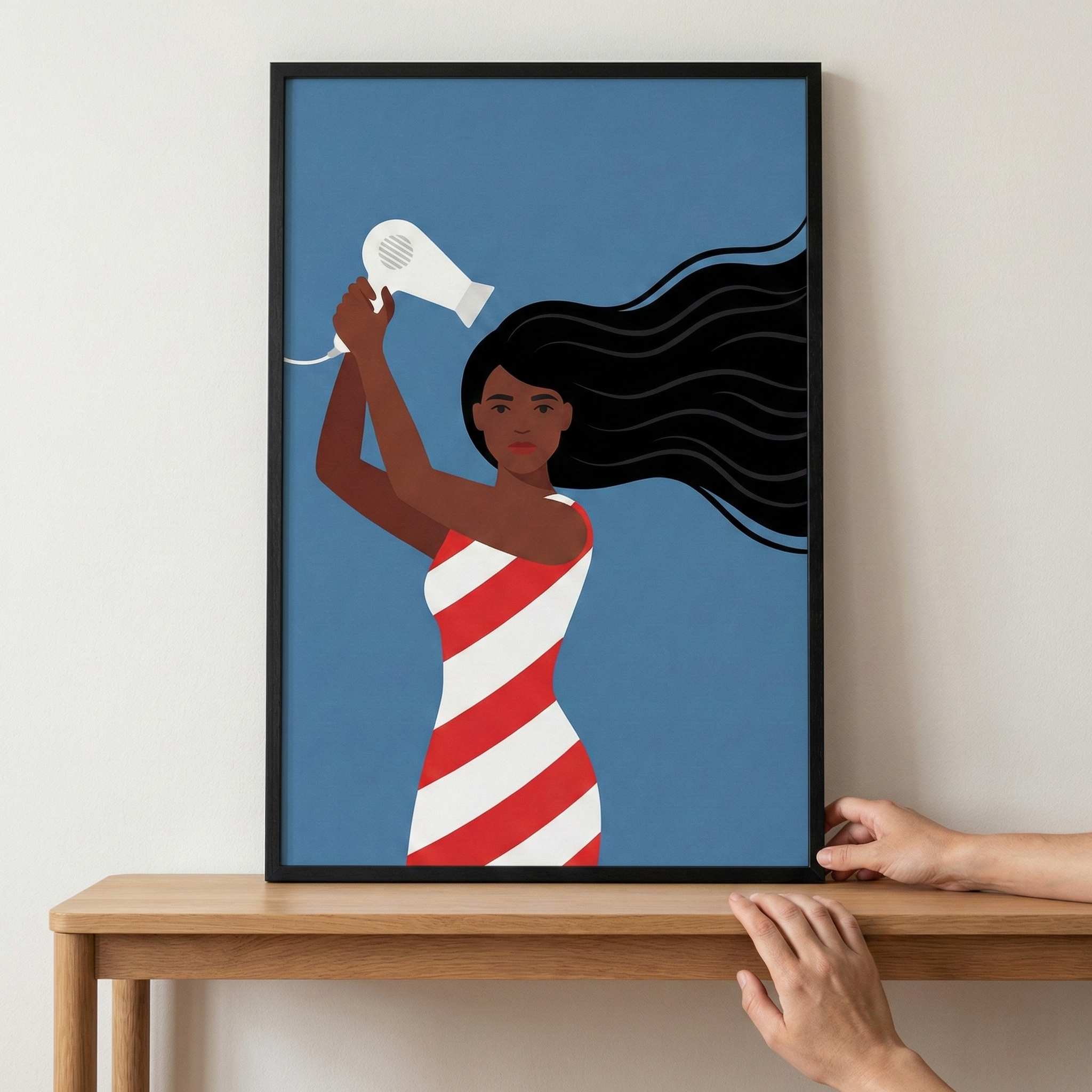 A minimalist graphic illustration is presented in a simple black frame, resting on a light wooden surface. The artwork features a woman with dark skin and exceptionally long, flowing black hair. She is captured from the waist up, holding a white hair dryer towards her hair with raised arms. She wears a sleeveless dress with bold diagonal red and white stripes. The background is a solid, medium blue. A person's hands are seen at the bottom and side, adjusting the position of the frame.