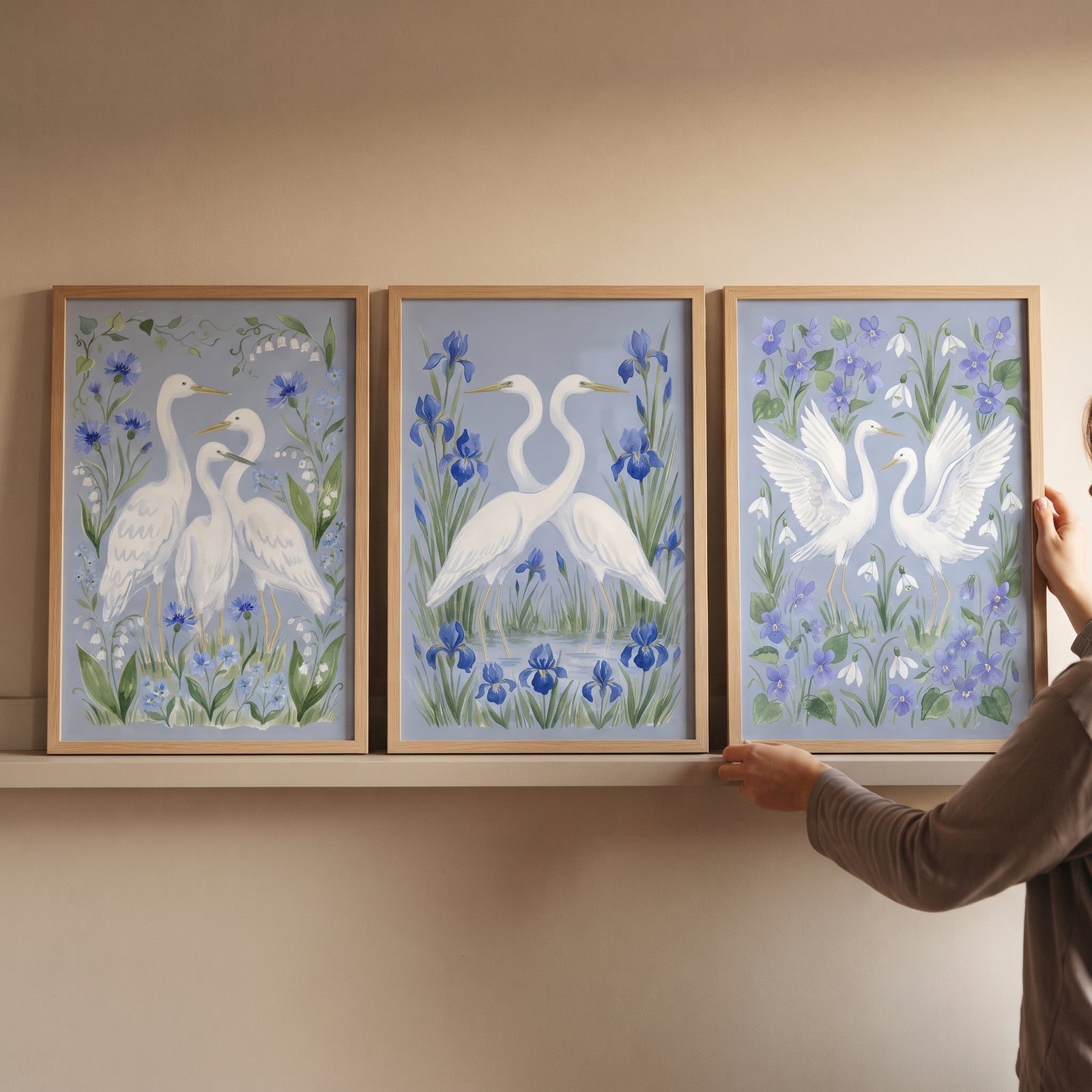 Hands adjust a blooming blue white egret bird wall art set of 3 featuring white birds and flowers in wood frames.