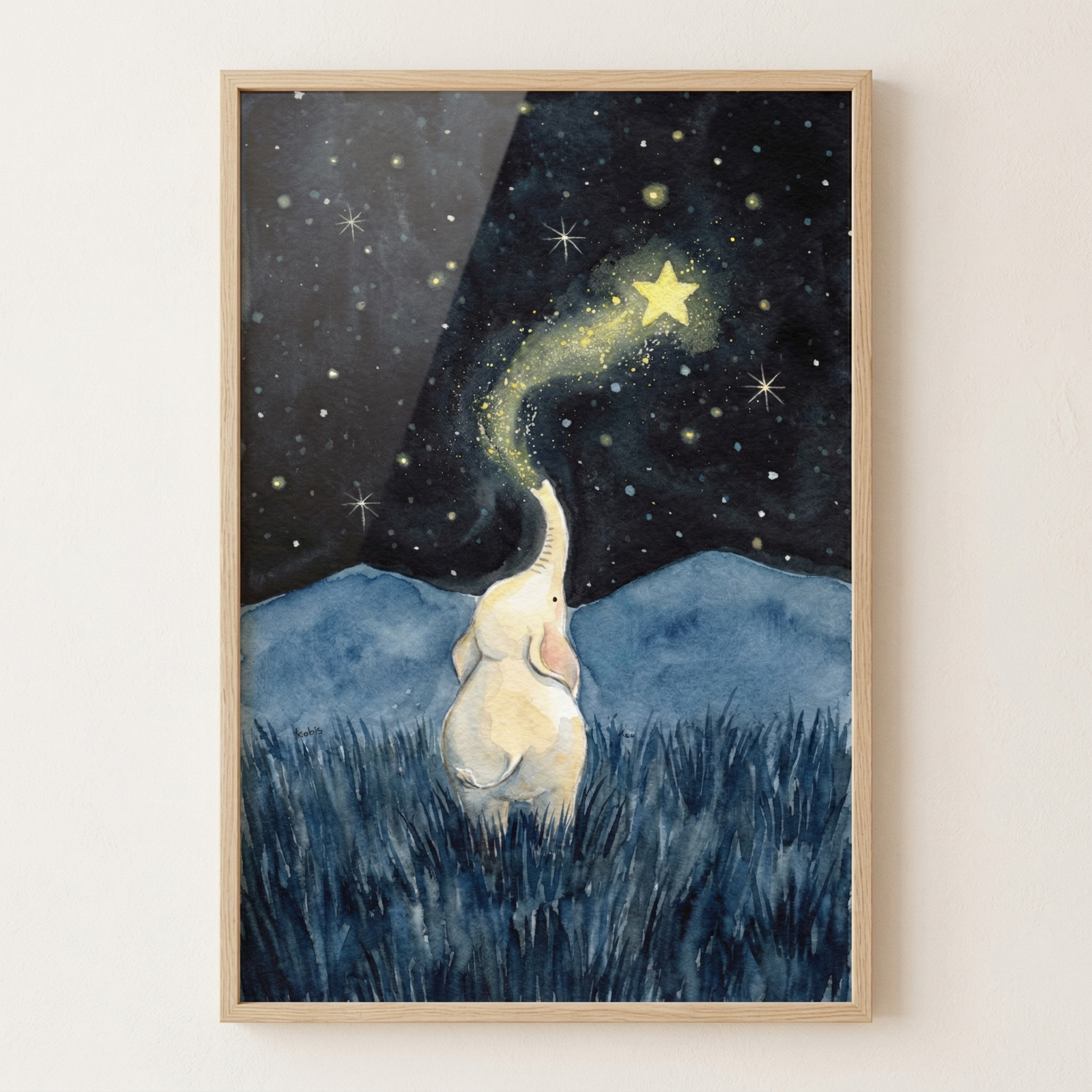 A framed piece of watercolor art depicts a small, cream-colored elephant from behind, standing in a field of dark blue grass. The elephant’s trunk is raised toward a black night sky filled with stars. A glowing trail of golden light and sparkles flows from the trunk up to a bright yellow star. In the distance, soft blue mountain silhouettes are visible against the starry sky. The work has a whimsical, dreamlike quality with soft textures.
