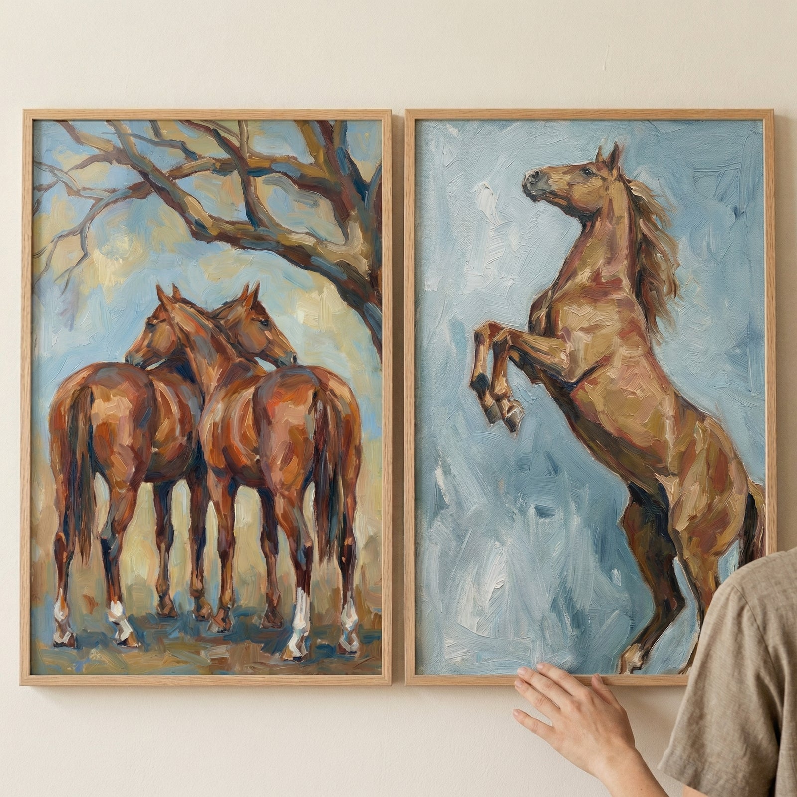 Two framed textured horse paintings displayed side by side, one showing two brown horses standing close together beneath tree branches and the other showing a rearing horse against a soft blue background. A hand adjusts the frame, suggesting artwork styling in a warm natural interior.