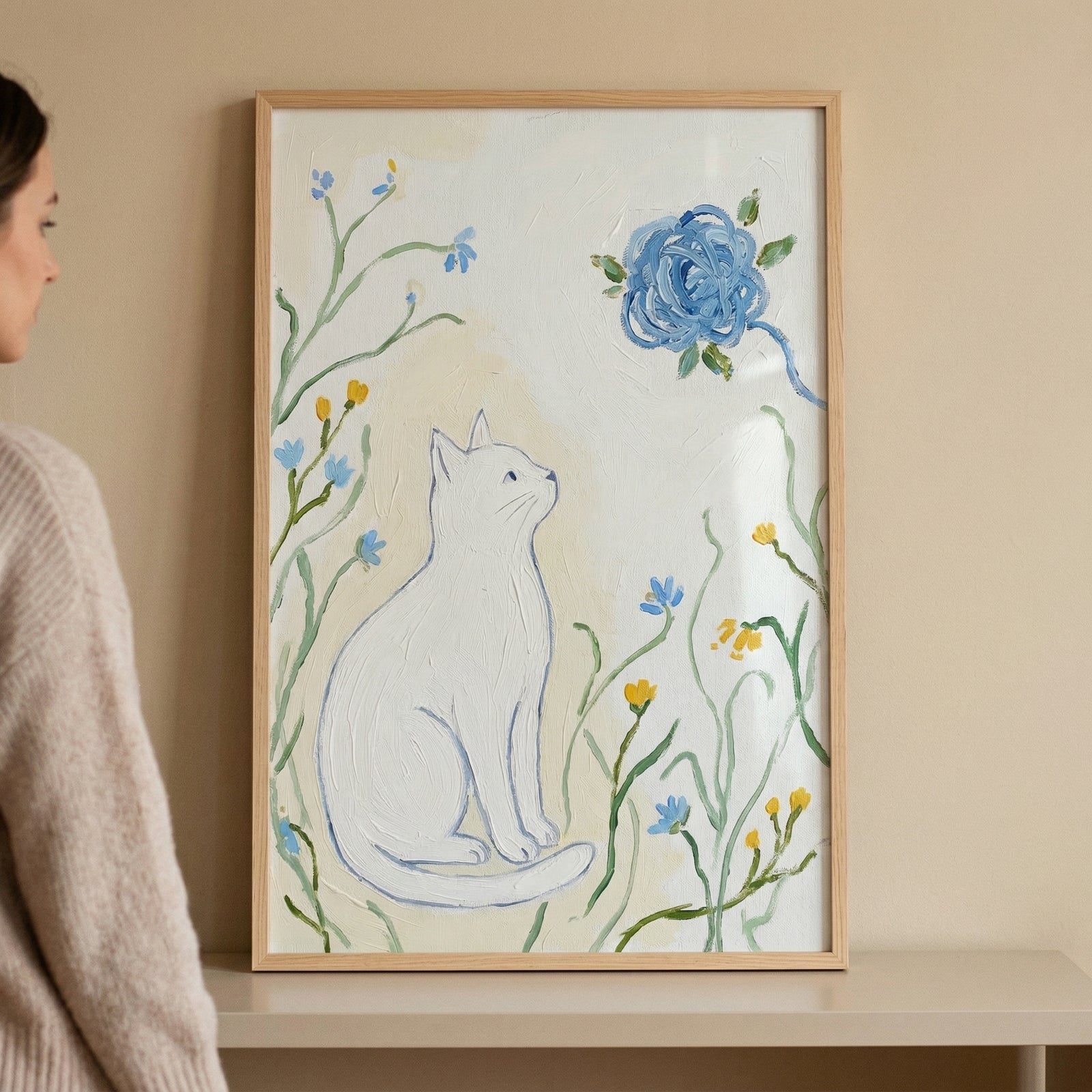 A vertical framed illustration hangs on a neutral wall. In the center, a white cat sits in profile, looking up toward a large, vibrant blue flower. The cat is surrounded by thin green stems with small blue and yellow blossoms. The piece has a thick, textured quality with visible brushwork on a cream-colored background. A person's profile is partially visible on the left side, gazing at the artwork.