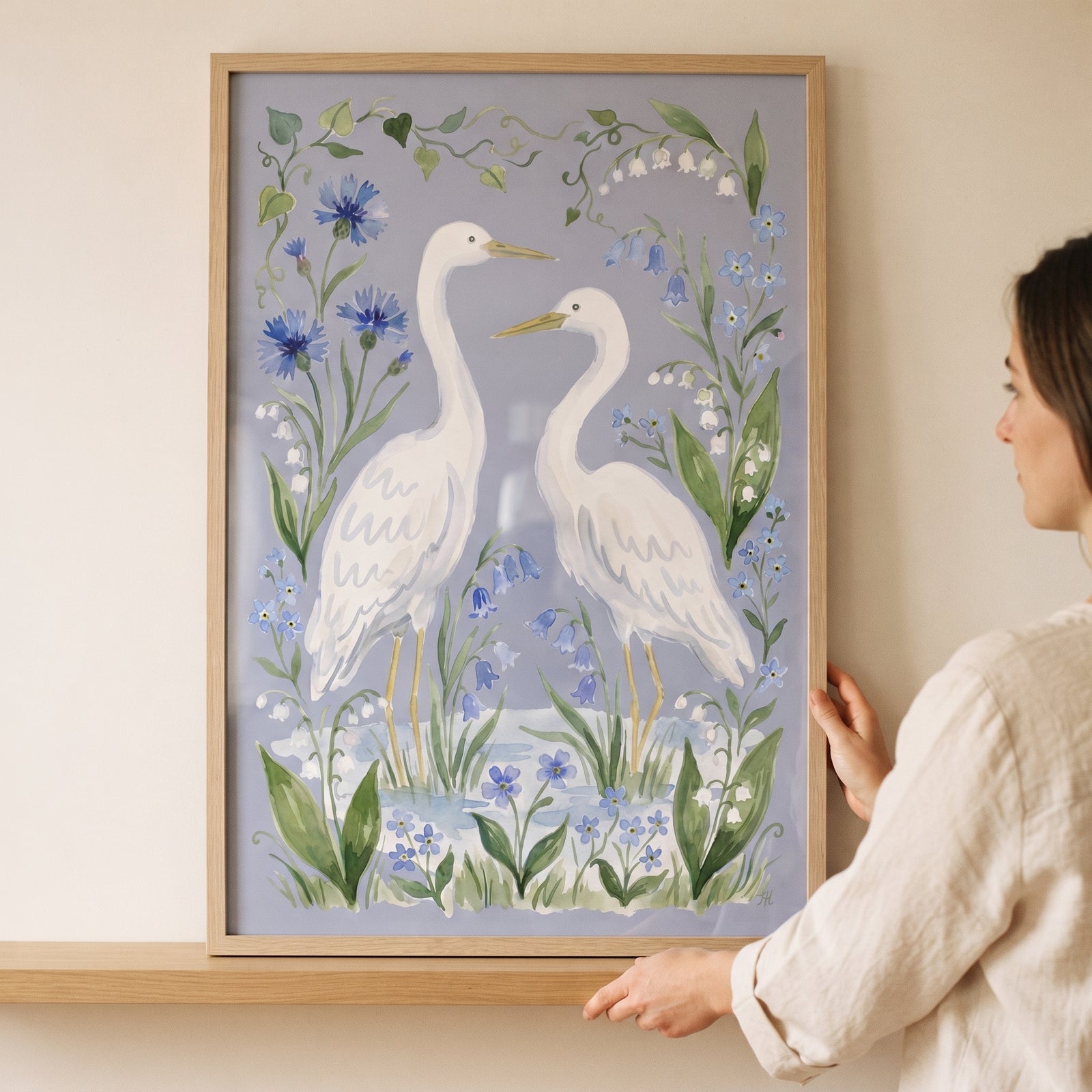 A person holds a framed blue botanical white egret bird wall art featuring two white birds and blue flowers.