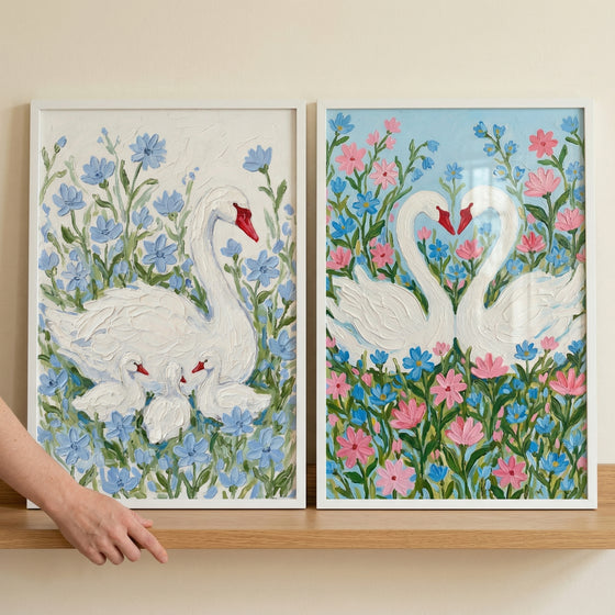Two framed textured paintings of swans among flowers, one showing a swan with two cygnets nestled in soft blue blooms and the other showing two swans forming a heart shape with their necks surrounded by pink and blue flowers. A hand points toward the artwork, suggesting placement or styling in a gentle home setting.