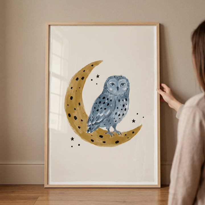 Framed blue crescent moon owl wall art shows a spotted blue owl on a golden moon being held against a neutral wall.