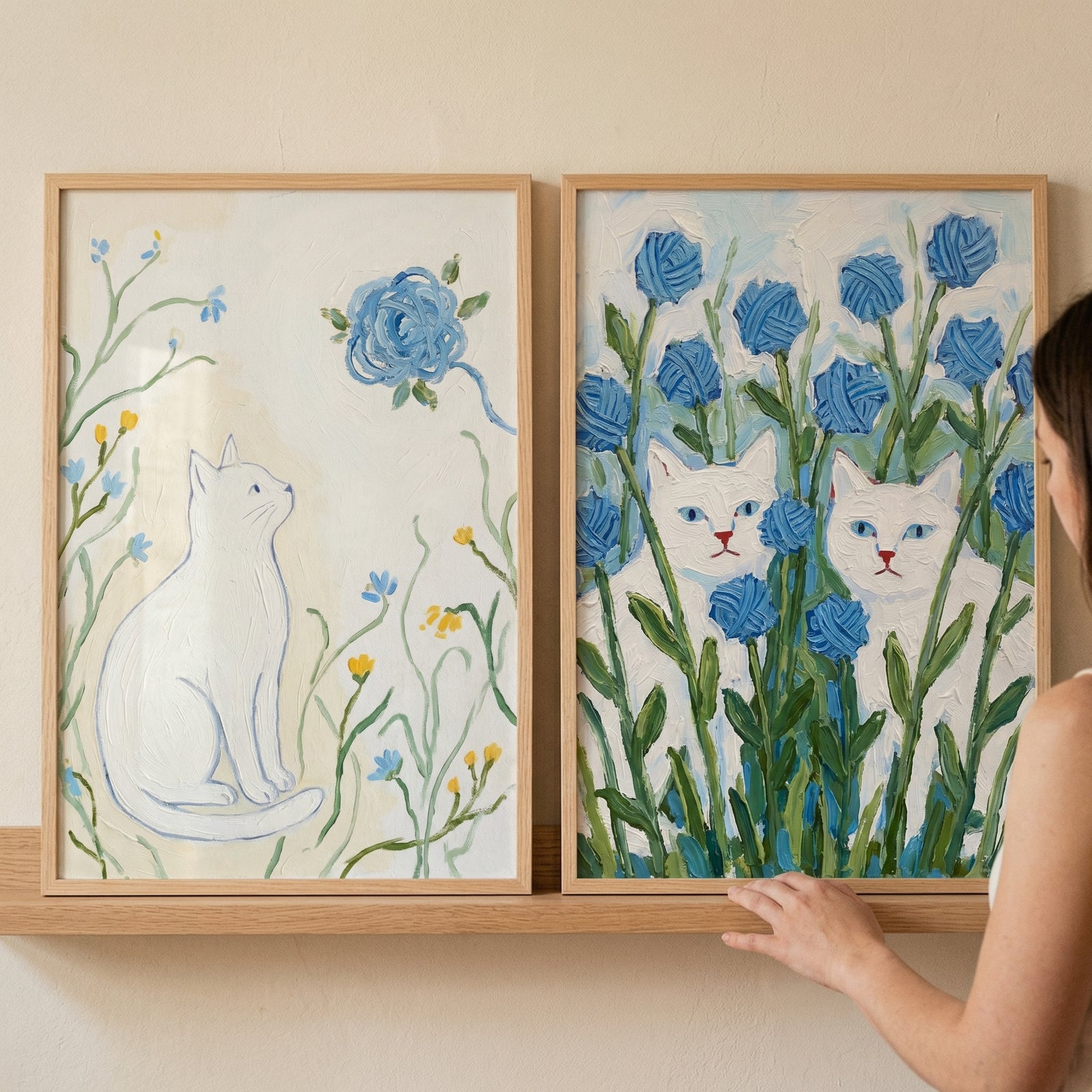 Resting on a wooden shelf against a beige wall, a person's hand adjusts one of two light wood-framed, textured paintings. The artwork features white cats and blue flowers: a single cat looking up at a large blue blossom among delicate stems on the left, and two cats peeking through a dense patch of tall blue flowers on the right.
