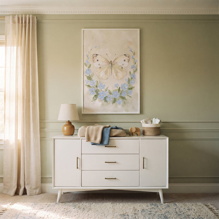 White dresser sits under blue floral wreath butterfly wall art on a sage green wall with cream curtains and a small lamp.