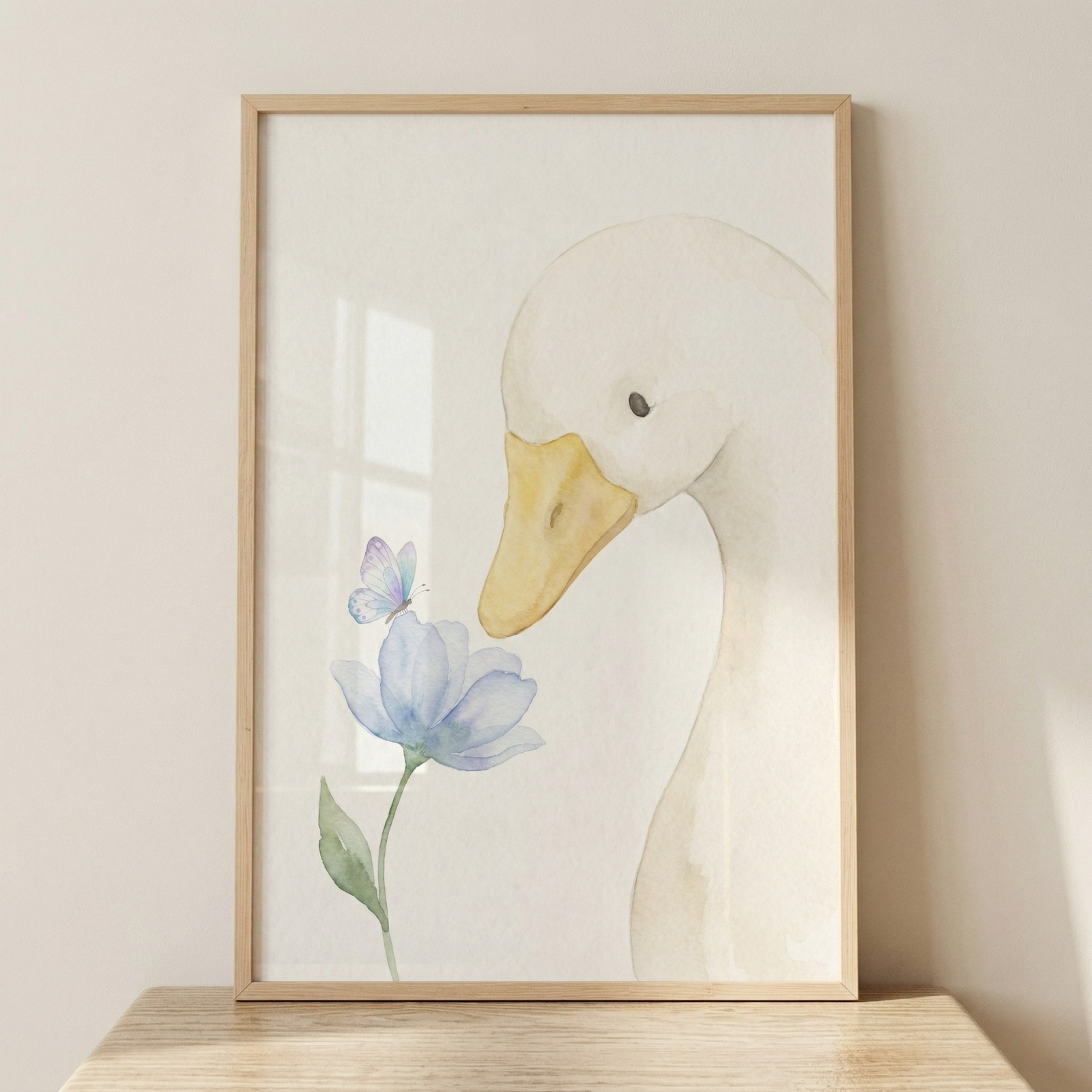 A framed watercolor illustration depicts a gentle white goose looking down at a delicate blue flower. The goose has a soft, rounded head, a small dark eye, and a pale yellow beak. On the edge of one of the flower's light blue petals sits a tiny, pale purple butterfly. The artwork is rendered with soft edges and a minimalist feel on an off-white background. It is displayed in a light-colored wooden frame sitting on a wooden surface against a neutral wall.