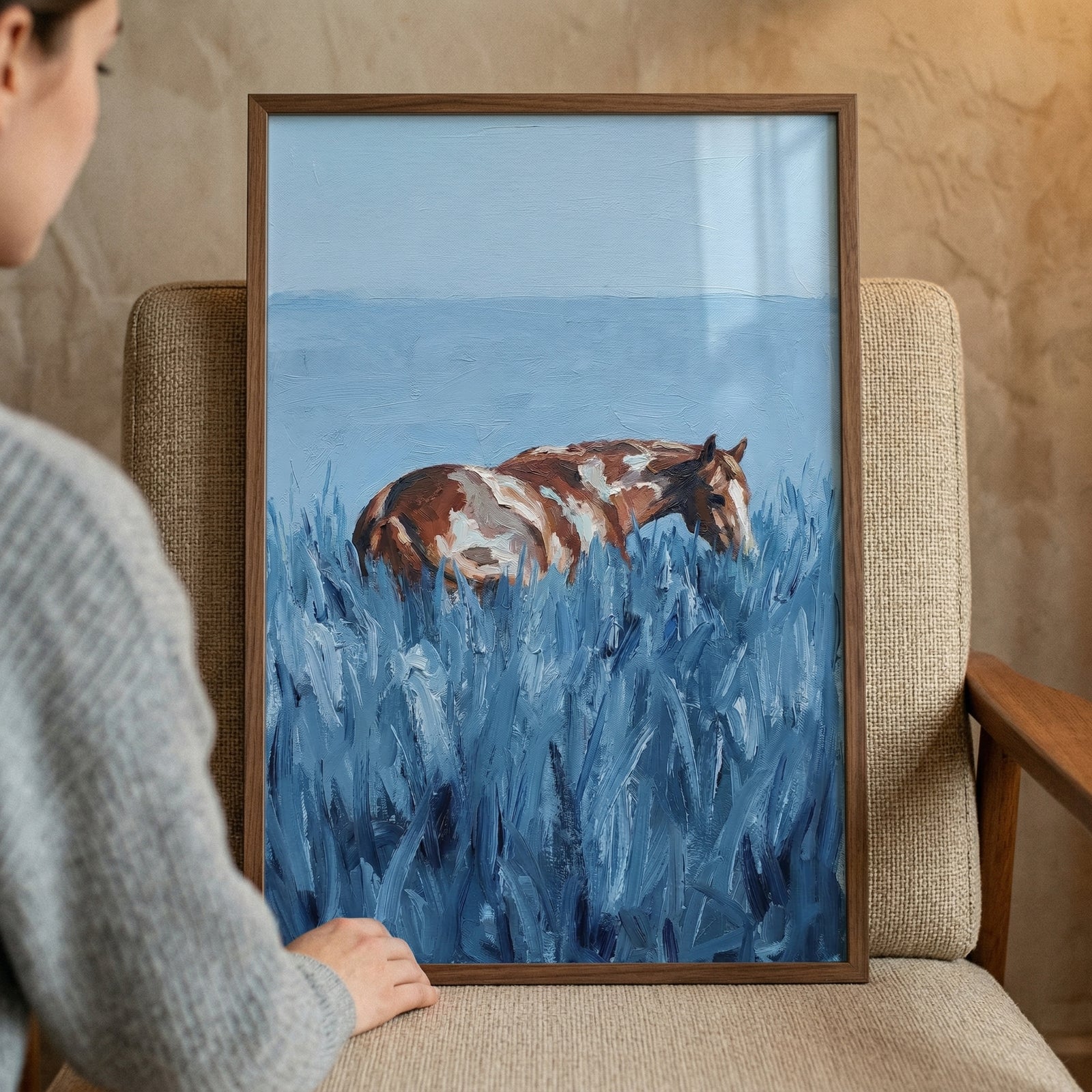 An expressive piece of art framed in wood depicts a brown and white pinto horse standing in a field of thick, blue-toned grass. The horse's coat is rendered in patches of earthy brown and white, while the surrounding vegetation is composed of heavy, vertical brushstrokes in various shades of blue, creating a sense of depth and texture. 