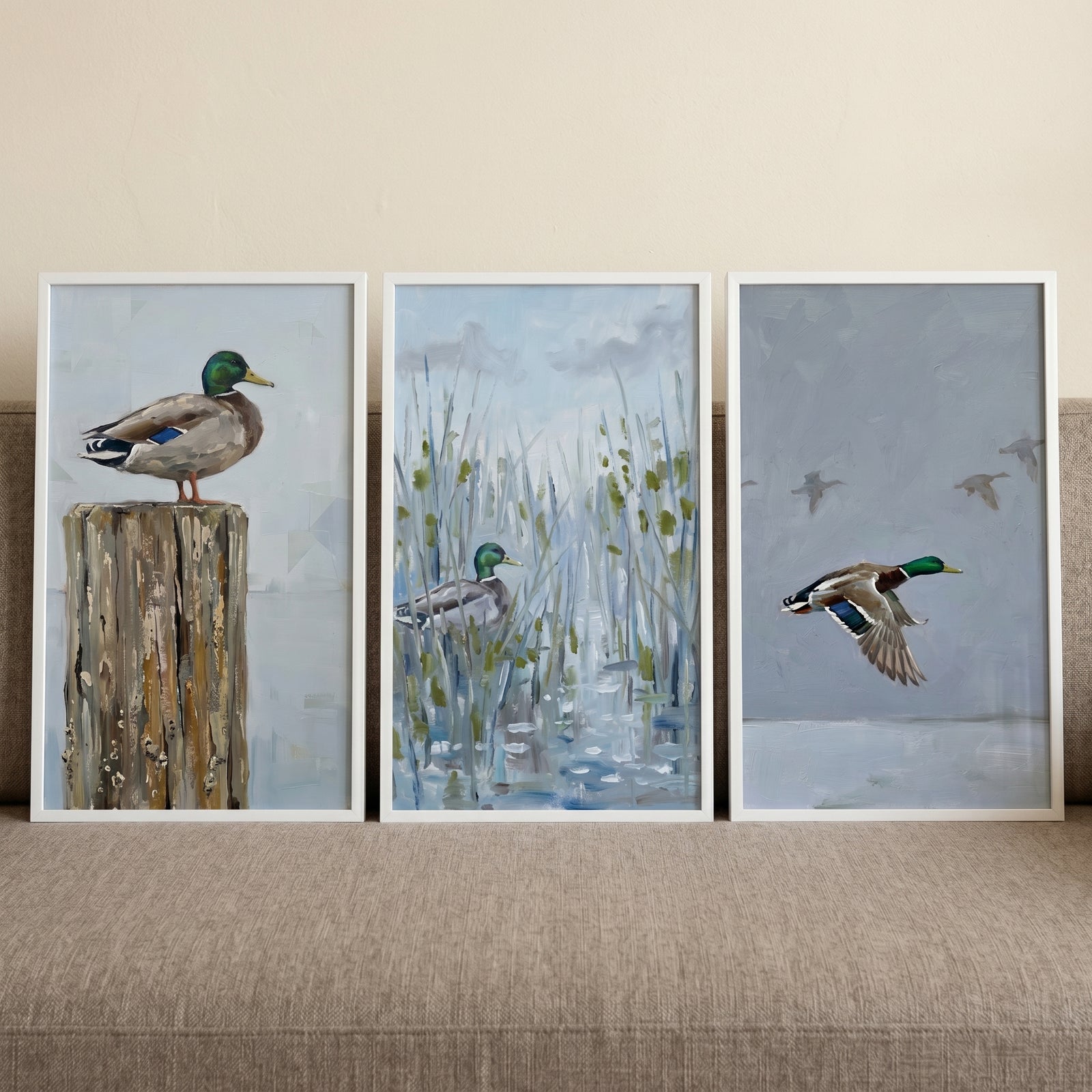 Blue grey mallard duck wall art set of 3 shows a mallard on a post, among reeds, and in flight against a grey sky.