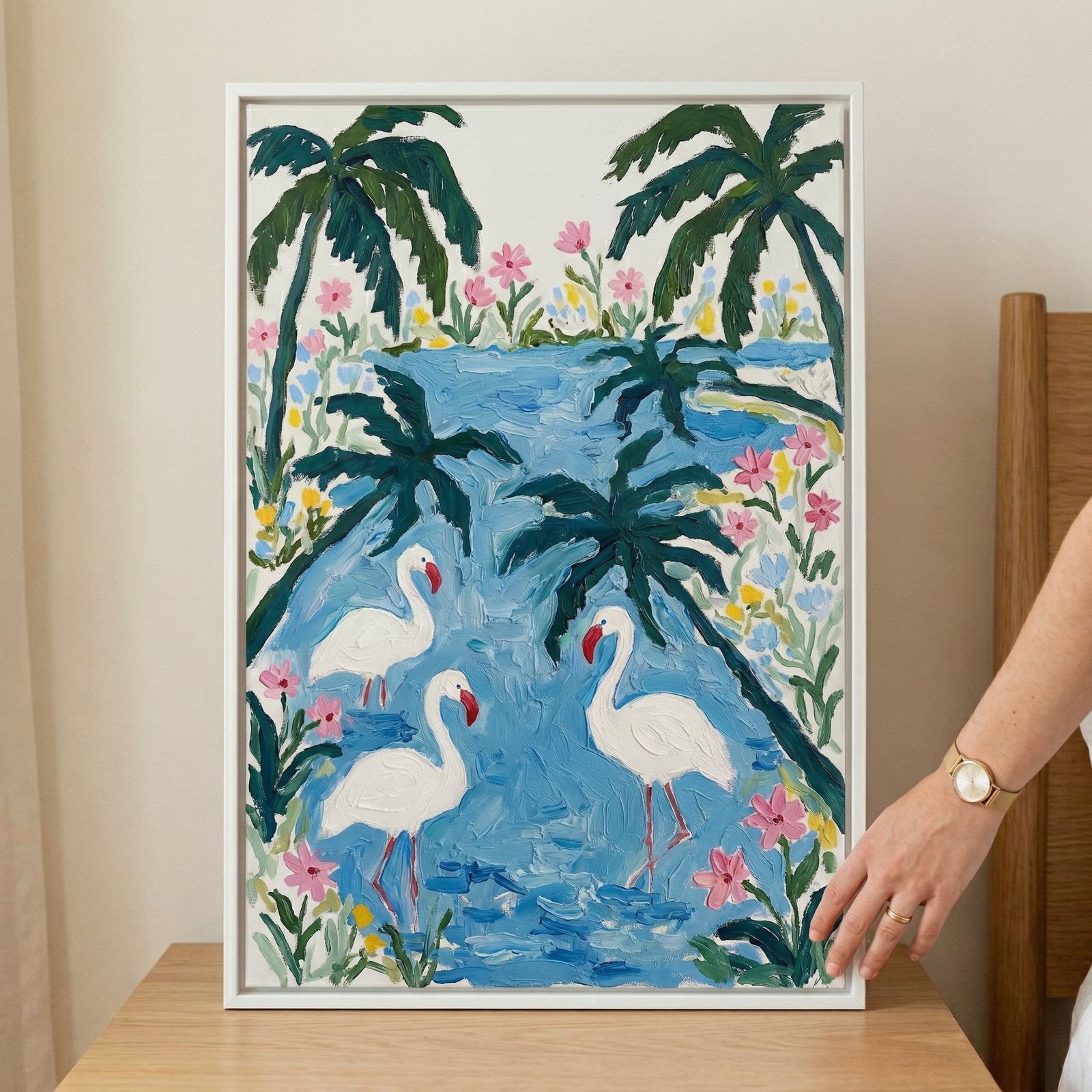 Framed painting of three white birds standing in blue water surrounded by pink flowers and palm trees with a waterfall in the background. A hand rests on the frame showing the scale and adding a styled lifestyle context.