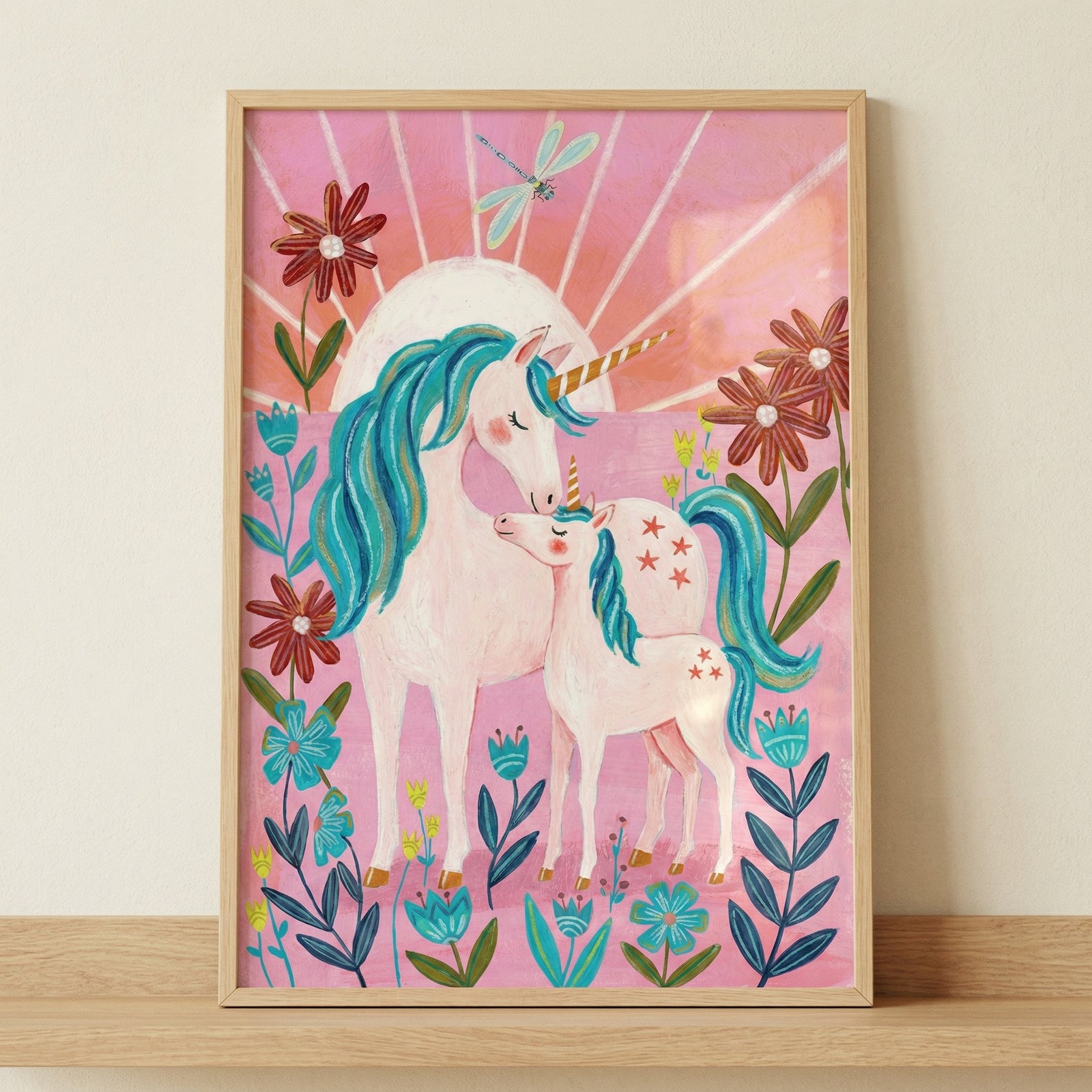 This framed illustration features a whimsical scene of a large white unicorn and its foal standing amidst a field of vibrant, stylized flowers. Both creatures have flowing teal manes and tails, and golden horns. The background is a warm pink, decorated with a bright sunburst and a delicate teal dragonfly. The artwork, with its folk-art-inspired style and playful colors, is displayed in a simple light wood frame resting on a wooden surface against a plain wall.