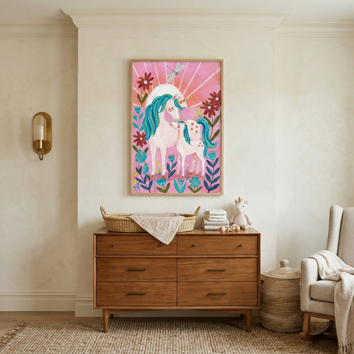 Framed illustration of a unicorn and baby unicorn with teal manes surrounded by colorful flowers on a pink background, styled above a wooden dresser. The artwork adds a whimsical, storybook feel to the nursery alongside soft textiles, a woven basket, and a cozy chair.