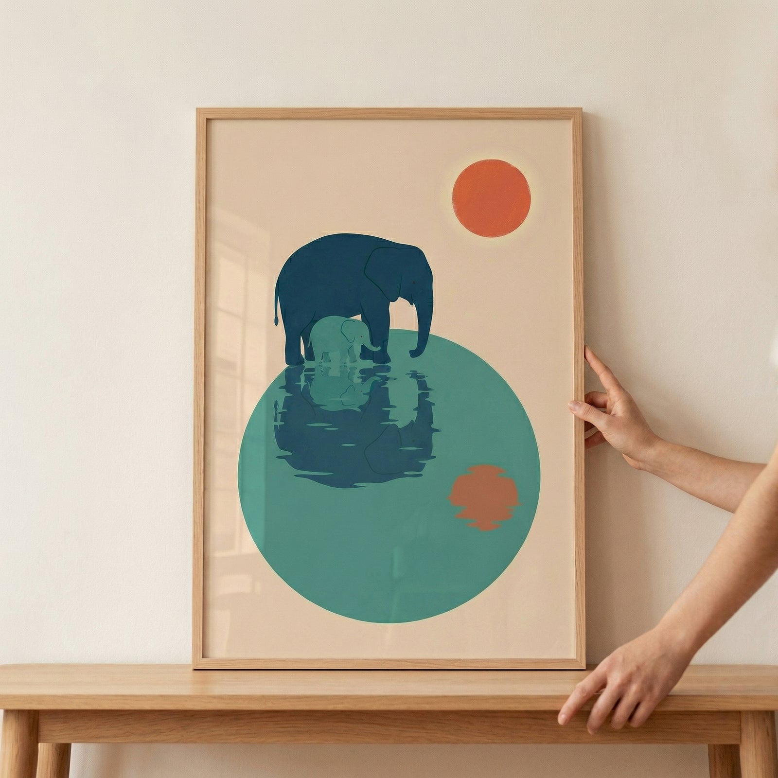 A framed minimalist illustration shows a large blue elephant and a smaller baby elephant standing in water with their reflections below and a warm orange sun above. A hand holds the frame in place on a wooden shelf, highlighting it as a styled nursery art piece.