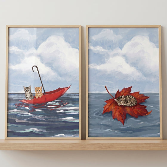 Blue ocean floating cat wall art set of 2 featuring kittens in a red umbrella and one asleep on a red leaf.
